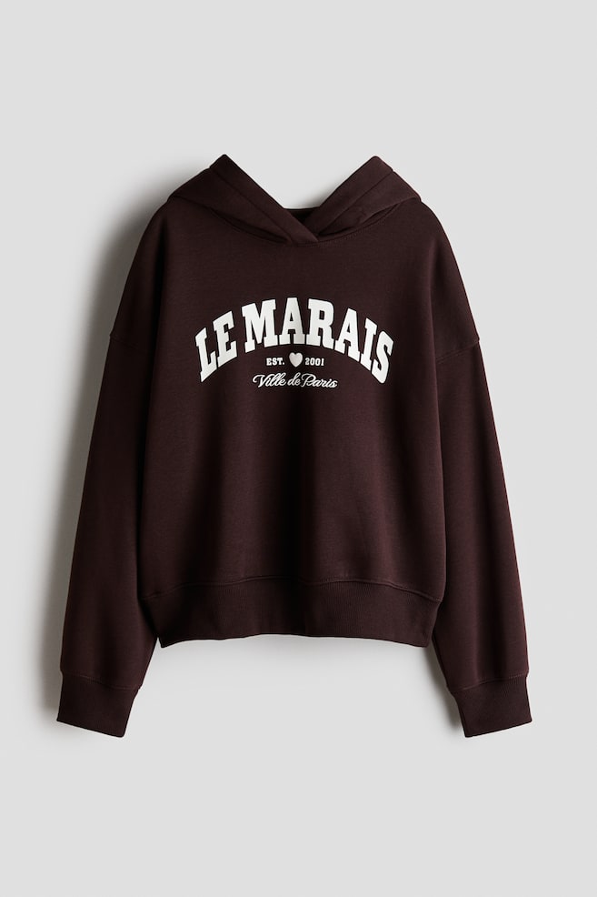 A dark brown fleece hoodie is shown from the front, featuring a crossover wrap hood, dropped shoulders, and long sleeves with ribbed cuffs and hem. The chest showcases a white varsity-style graphic print that reads "LE MARAIS" with "EST. 2001" and "Ville de Paris" written underneath. - 1