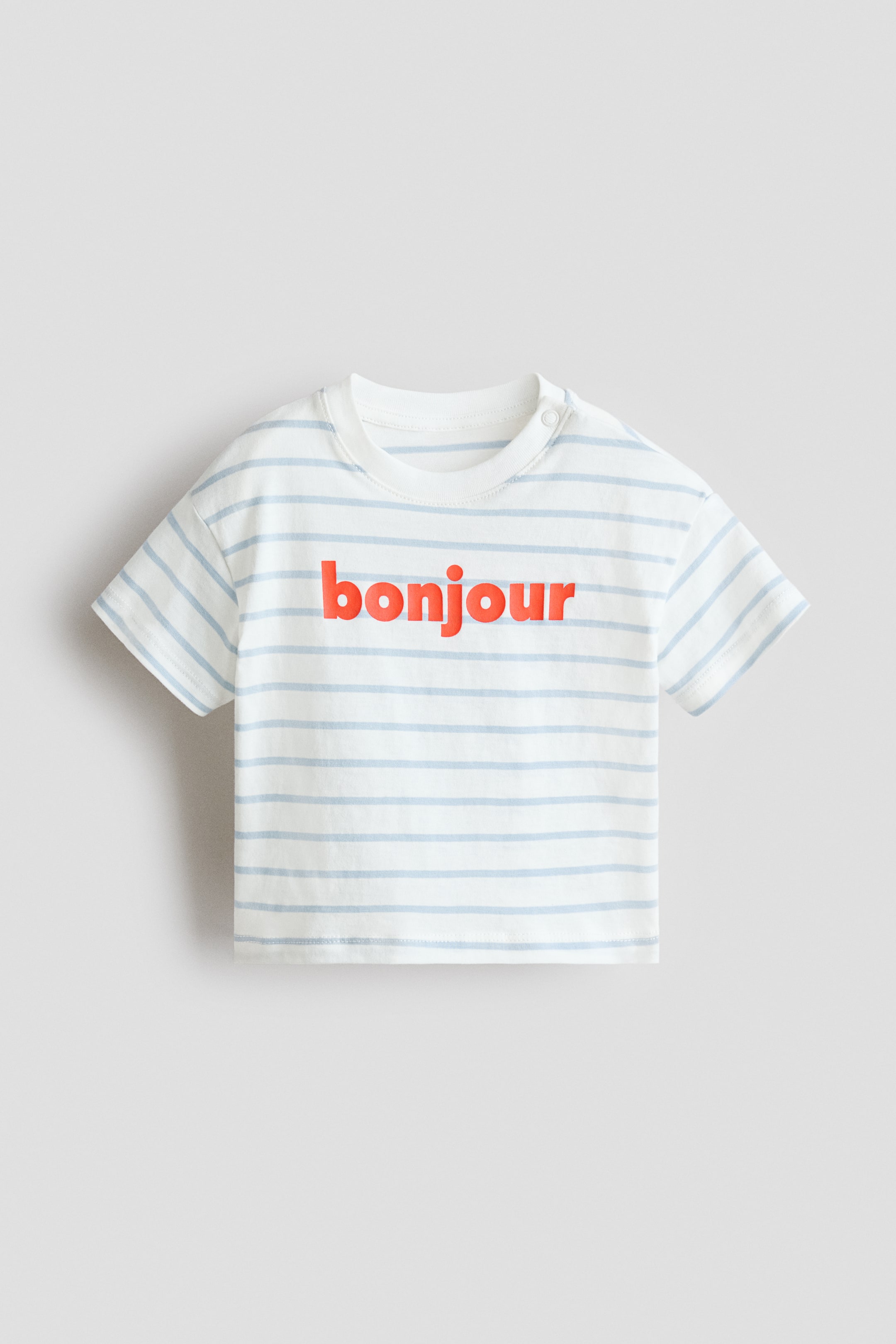 View larger image: A cotton T-shirt shown from the front, made from white fabric with light-blue horizontal stripes and the red lowercase word 'bonjour' printed across the chest. It features a crew neck, short sleeves and two snap buttons on the left shoulder.
