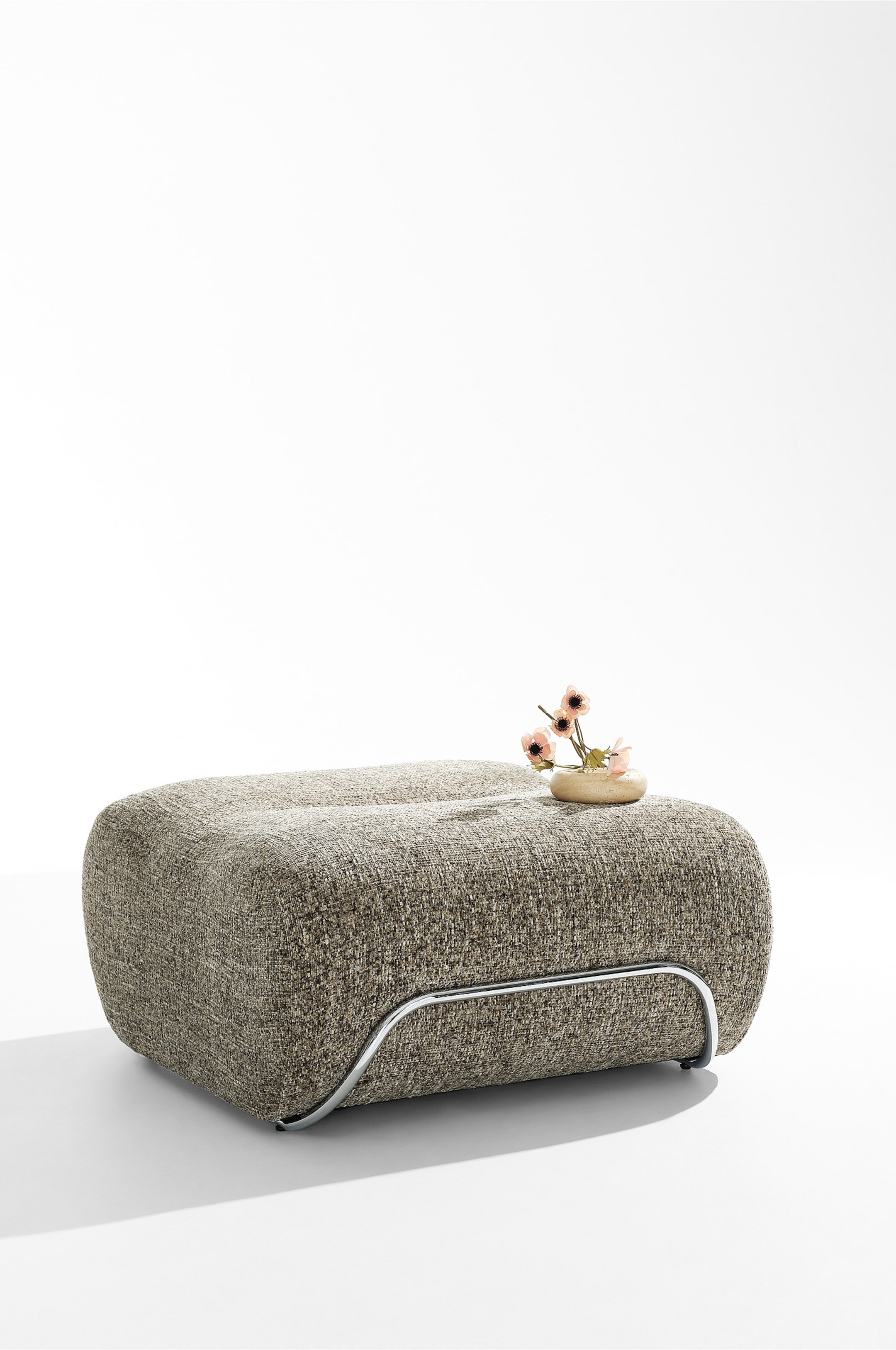 Visa större bild: Archie Pouf vilar i en trekvarts vinkel och framhäver sin lågprofilerade, avrundade rektangulära design klädd i ett texturerat, flerfärgat vävt tyg i brunt och beige. Den har en böjd, rörformad ram i krommetall längs den nedre kanten för stöd, med en liten dekorativ keramikskål med rosa blommor placerad på ovansidan.