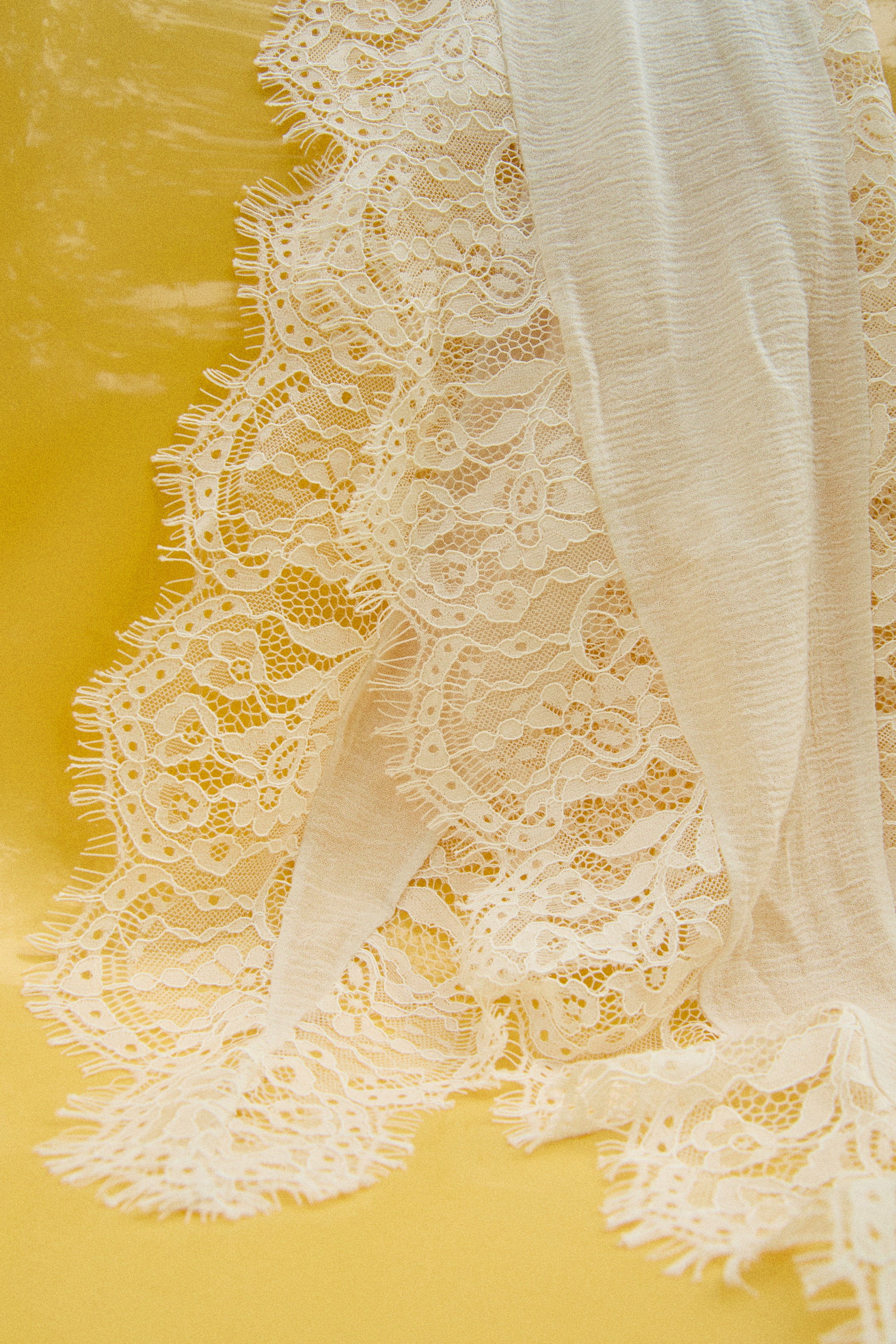 Agrandir l'image: Un tissu crème vaporeux et transparent, à la texture légèrement froissée, recouvre partiellement un tissu en dentelle de couleur crème, présentant de délicats motifs floraux et des bords festonnés agrémentés de franges effet cils.