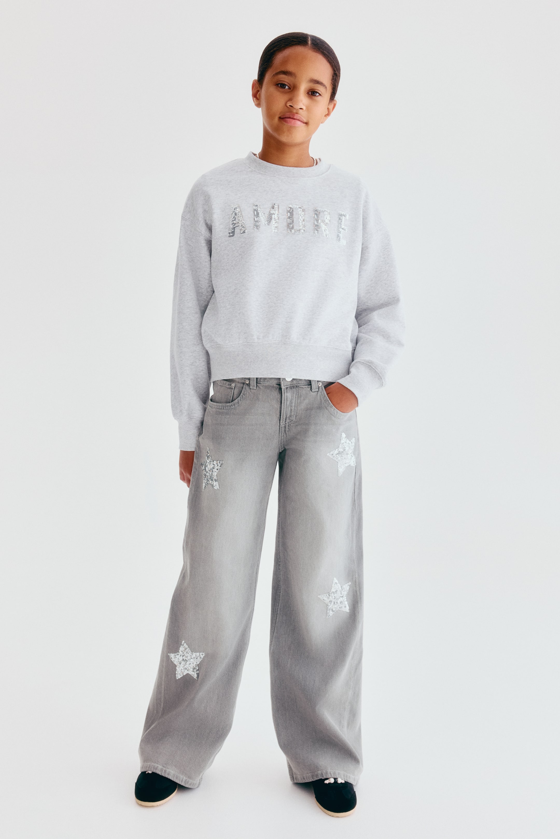 View larger image: A pair of wide-leg, grey cotton denim jeans is shown from the front, featuring a low-rise waist and five-pocket styling. The jeans are decorated with silver-sequined star patches scattered down the legs and are paired with a grey sweatshirt.