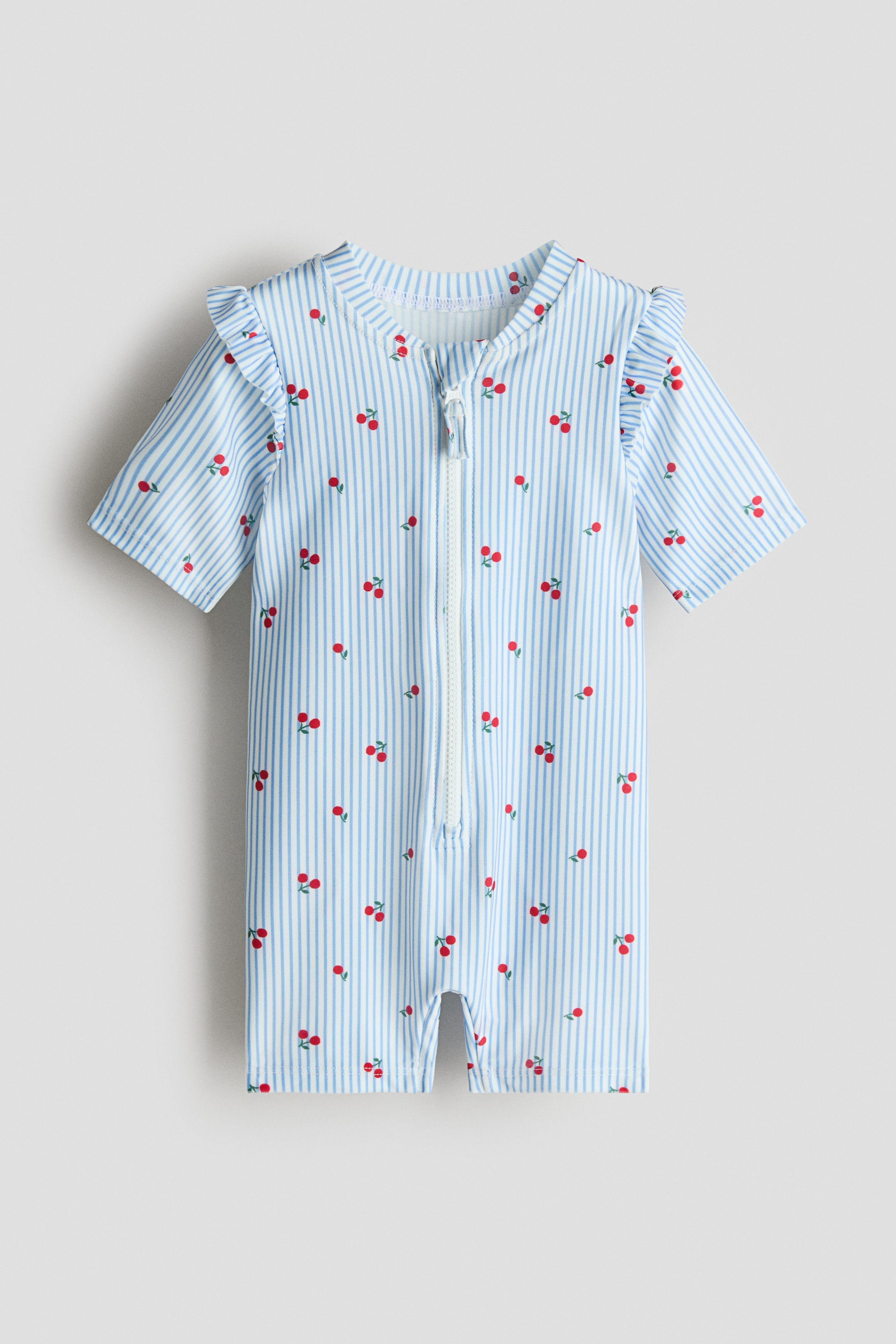 Grotere afbeelding bekijken: Een badpak toont de VOORKANT, met een lichtblauw-wit gestreept patroon met rode kersenmotieven. Het is ontworpen met een opstaande kraag, korte mouwen met ruches, korte pijpen en een doorlopende rits over de volledige lengte aan de voorkant.