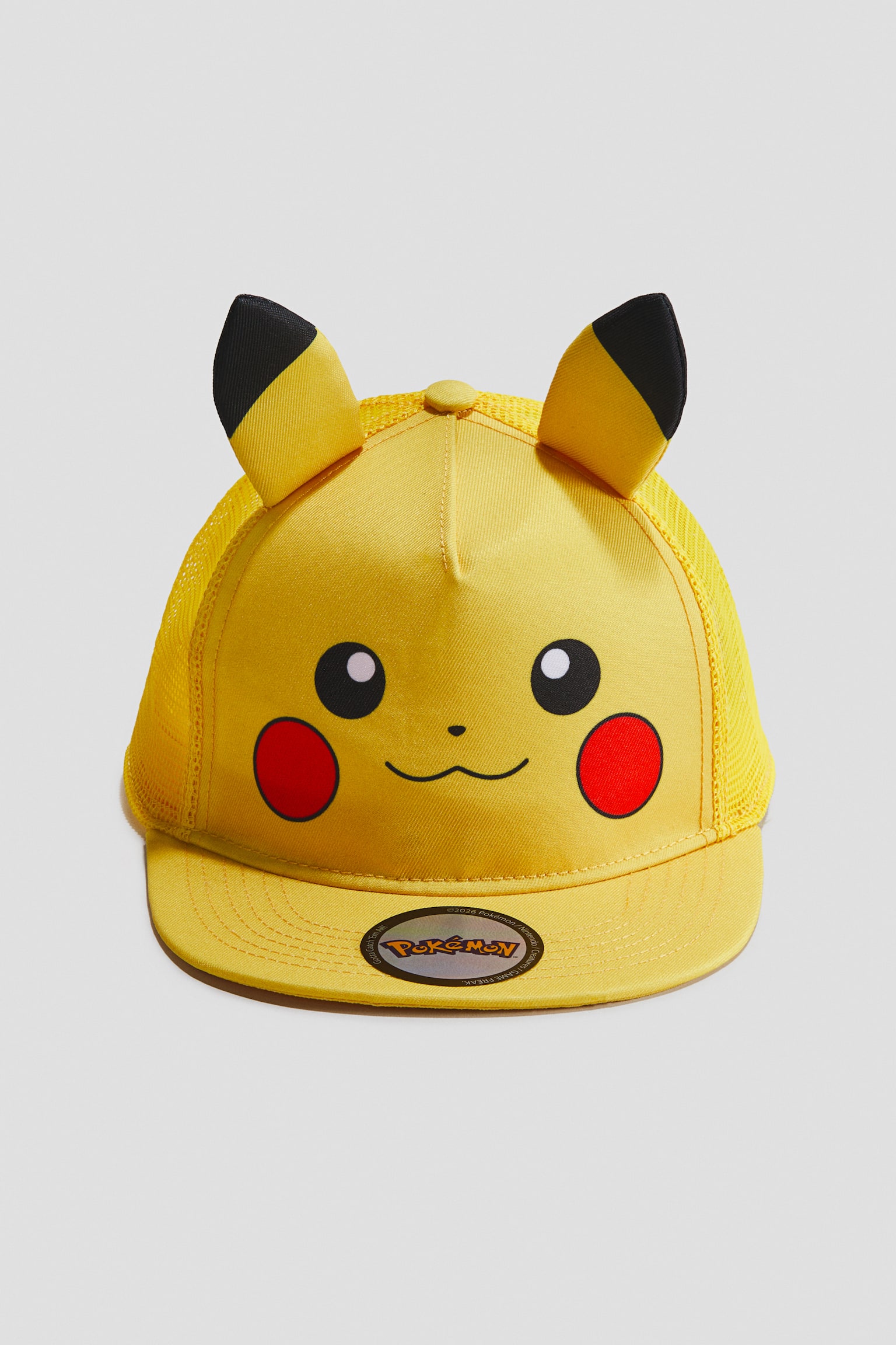 Chiếc mũ lưỡi trai màu vàng đang trình bày mặt trước của một khuôn mặt Pikachu trên tấm panel phía trước, nổi bật với đôi mắt đen, má tròn đỏ và một miệng nhỏ màu đen. Hai tai đệm màu vàng với đầu đen đứng thẳng trên đỉnh, trong khi mũ được thiết kế với phần lưới phía sau màu vàng và vành mũ phẳng màu vàng hiển thị nhãn dán logo Pokémon holographic.