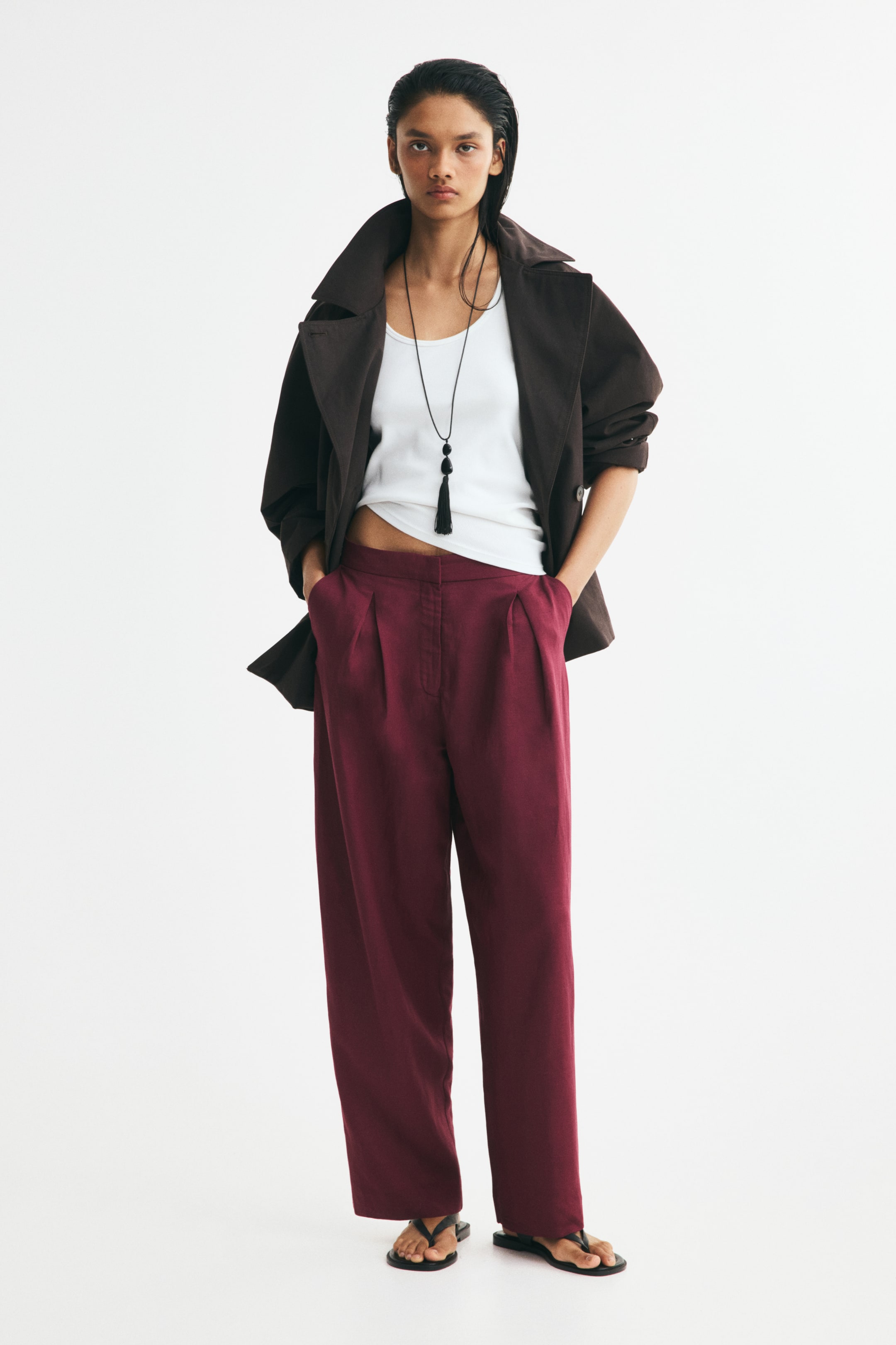 View larger image: Burgundy loose-fit pants featuring a high waist, front pleats, and a wide barrel-leg silhouette are being worn by the model, who is standing with hands tucked into the diagonal side pockets. The pants are styled with a tucked-in white tank top, an open dark brown jacket, and black flat sandals.