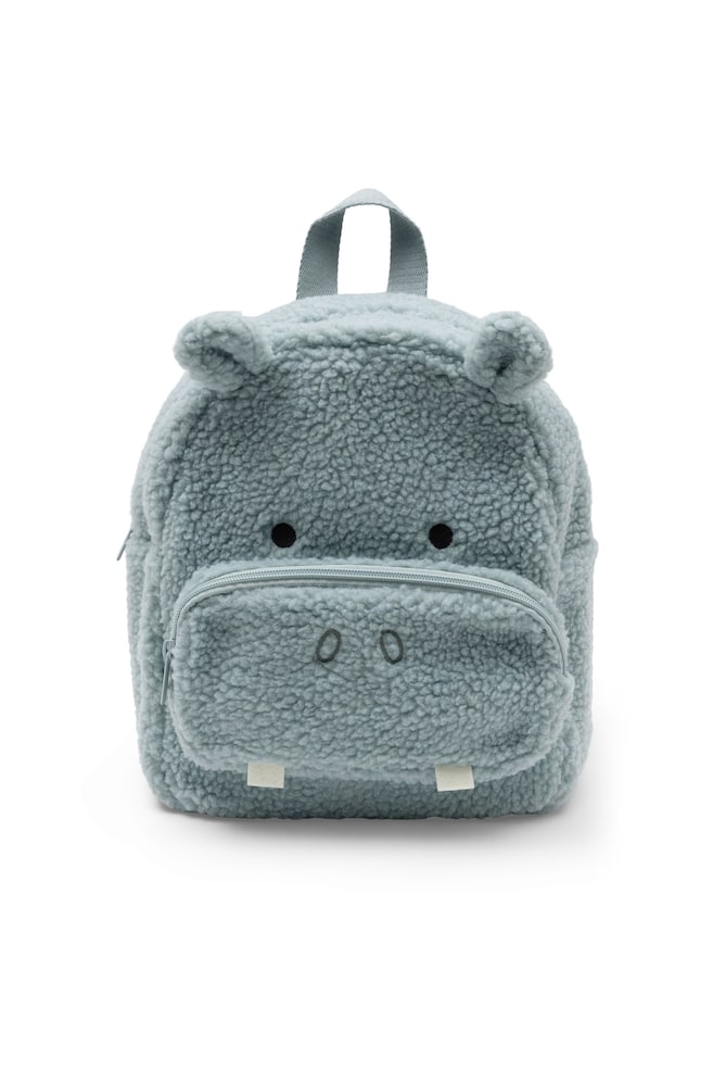 Un sac à dos pour enfant en polaire sherpa bleu-gris clair est présenté debout, de face, et conçu pour ressembler à un hippopotame. Le sac à dos comporte une poignée supérieure en sangle, de petites oreilles arrondies, des yeux noirs brodés, ainsi quâune poche frontale zippée agrémentée de narines brodées et de deux dents en feutre blanc. - 1