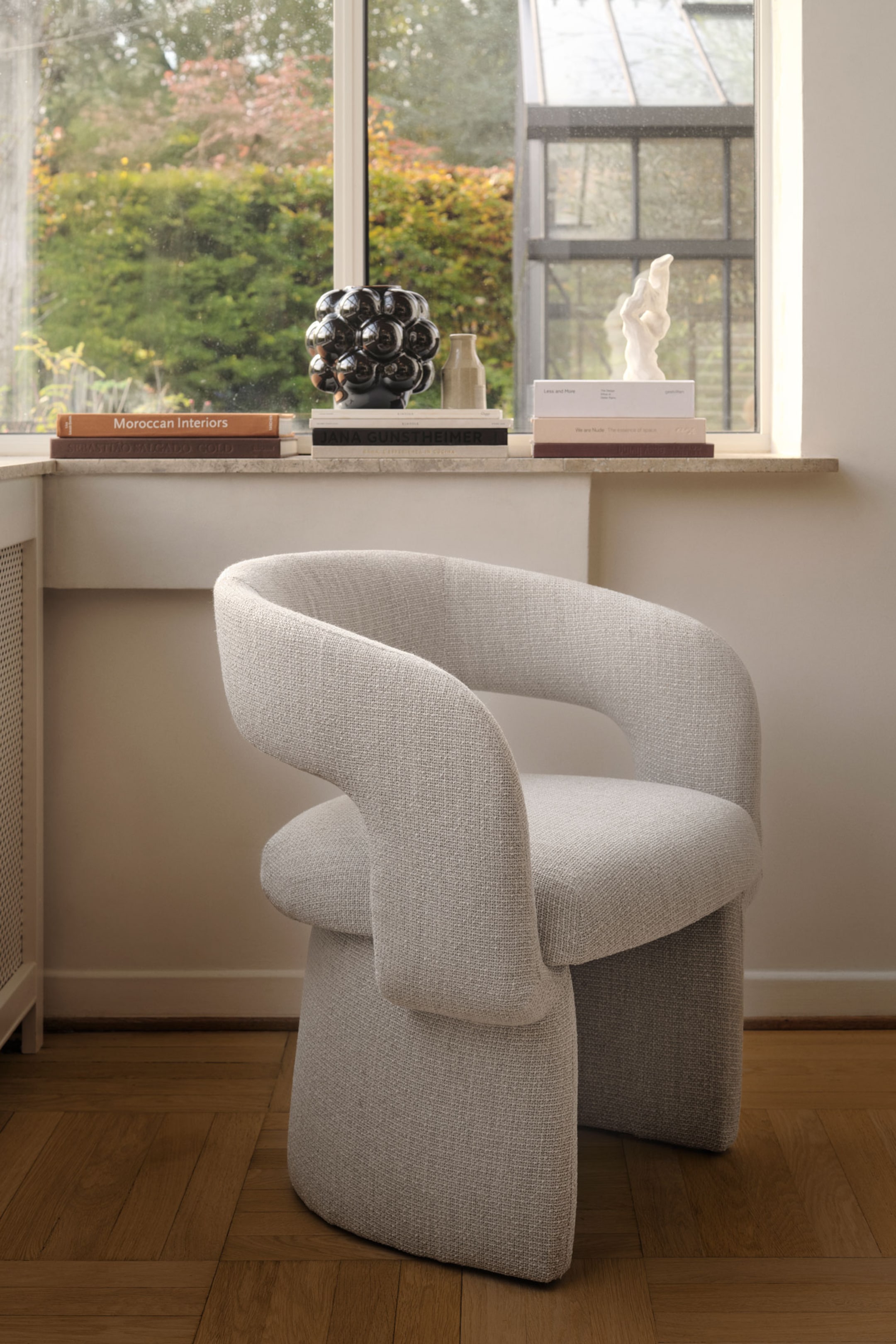 Grotere afbeelding bekijken: Een lichtgrijze, gestructureerde stoffen fauteuil staat in driekwartaanzicht naar rechts gericht, met een dikke, afgeronde zitkussen en een gebogen rugleuning die doorloopt en geïntegreerde armleuningen vormt. De stoel rust op twee brede, gebogen, blokvormige poten die bekleed zijn met dezelfde bijpassende stof.