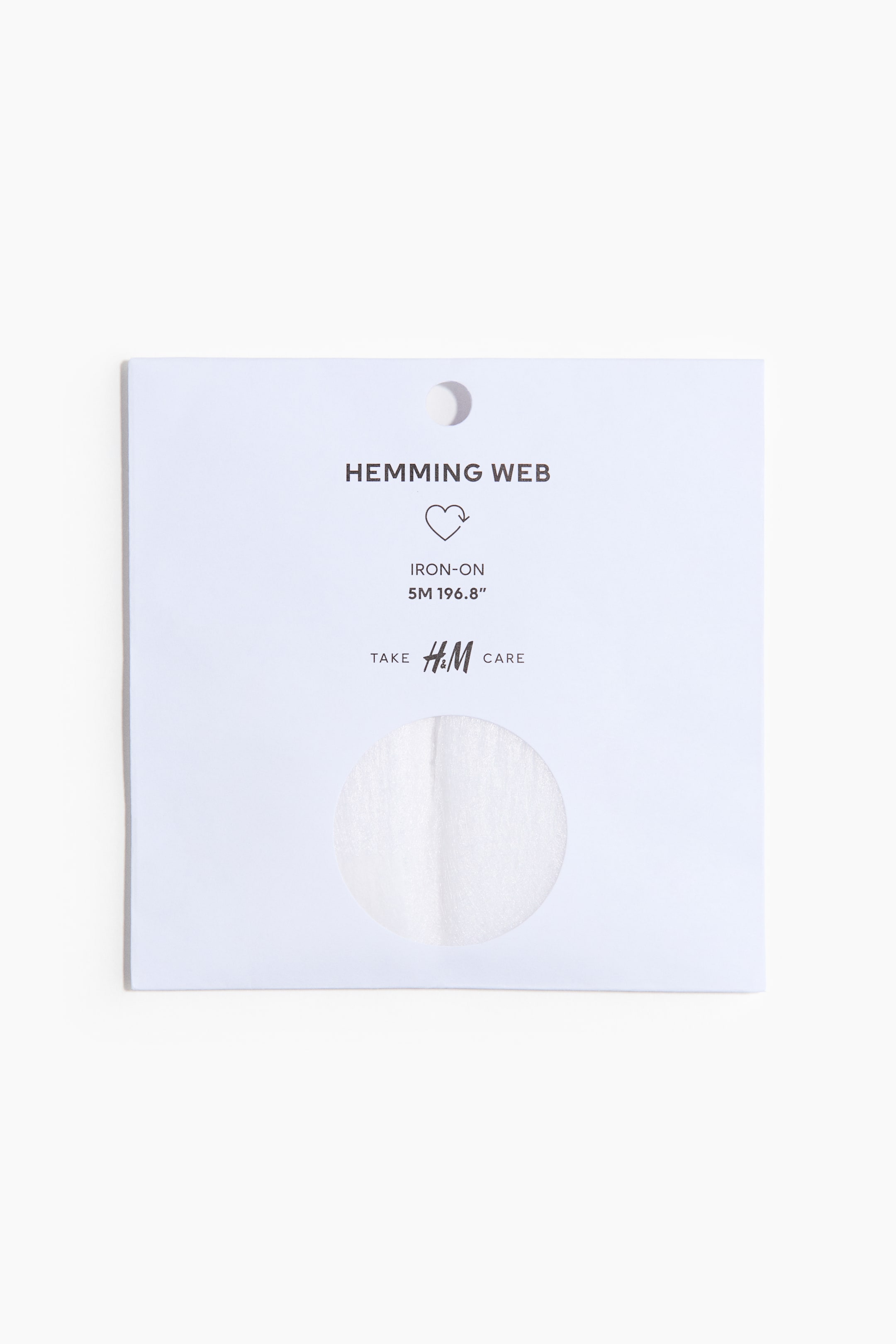 הצגת תמונה גדולה יותר: רצועת הדבקה לגימור להגהצה ארוזה בכרטיס מלבני לבן. החזית מציגה את הטקסטים "HEMMING WEB" ו-"IRON-ON", לצד המדידה "5M 196.8\"", סמל מיחזור בצורת לב וכתובית "TAKE H&M CARE" יחד עם לוגו H&M. חיתוך עגול בחזית חושף את רצועת ההדבקה הלבנה, חצי-שקופה, ונקב עגול קטן נראה בחלק העליון.