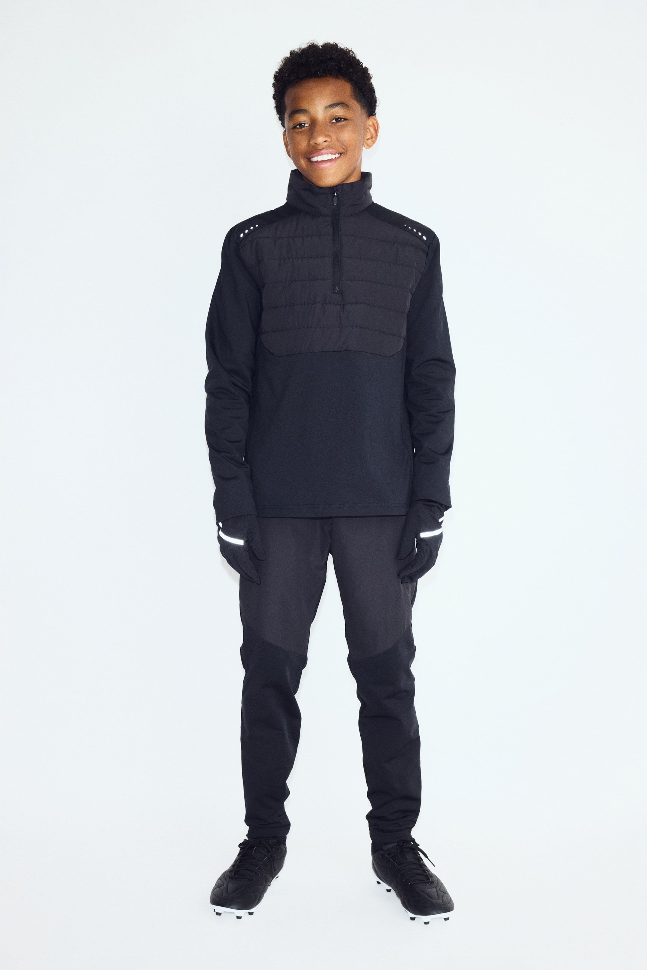 View larger image: A black athletic training outfit is shown from the front, featuring a long-sleeve half-zip top with a quilted chest panel and a high stand collar. The top is paired with slim-fit athletic pants featuring tonal paneling, black gloves with reflective details, and black soccer cleats.