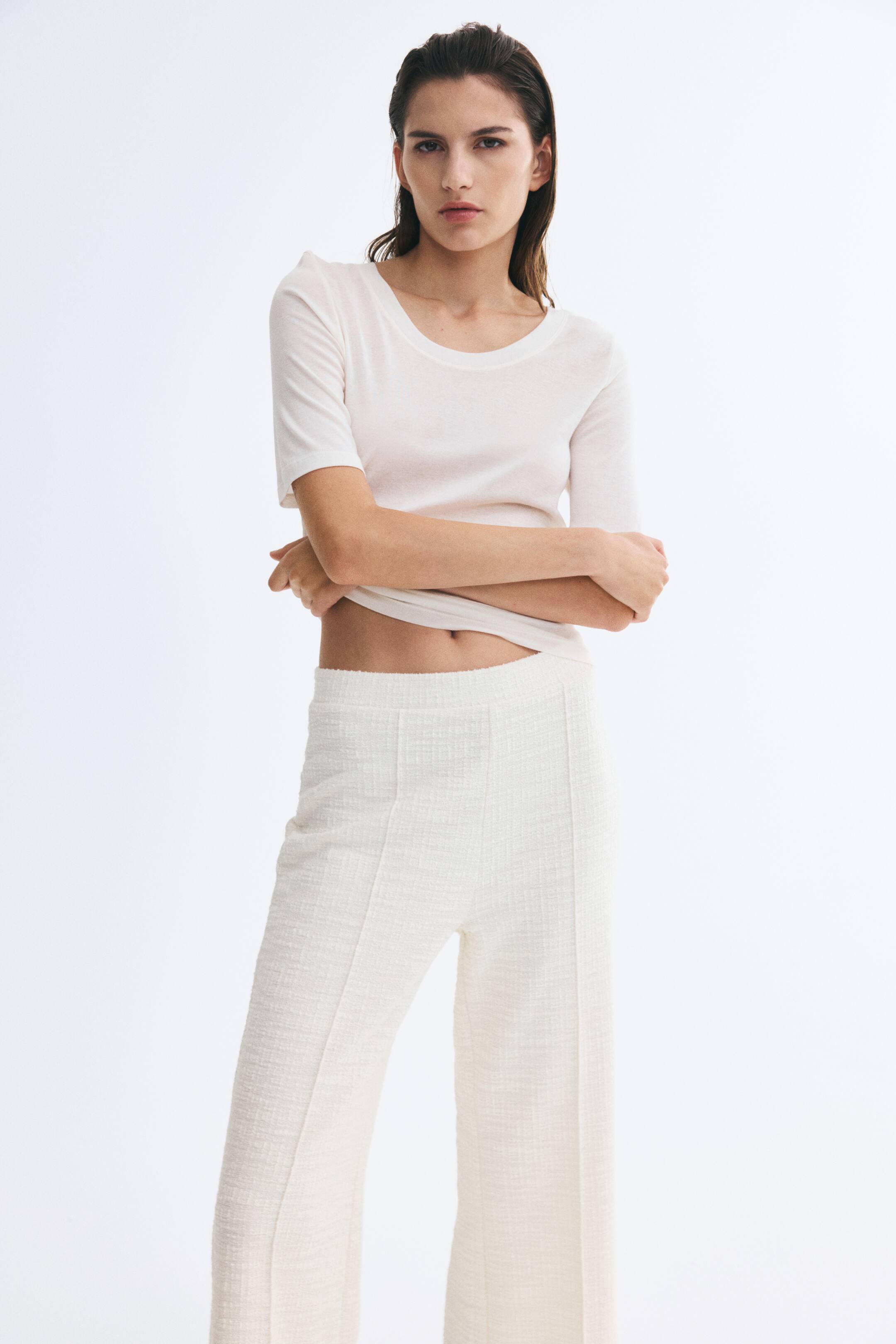 Ampliar la imagen: La modelo lleva puesta una playera color crema claro con cuello redondo, cruzando los brazos y levantando ligeramente el dobladillo. La playera se combina con un pantalón de pierna ancha en blanco roto con textura y detalle de costura central.