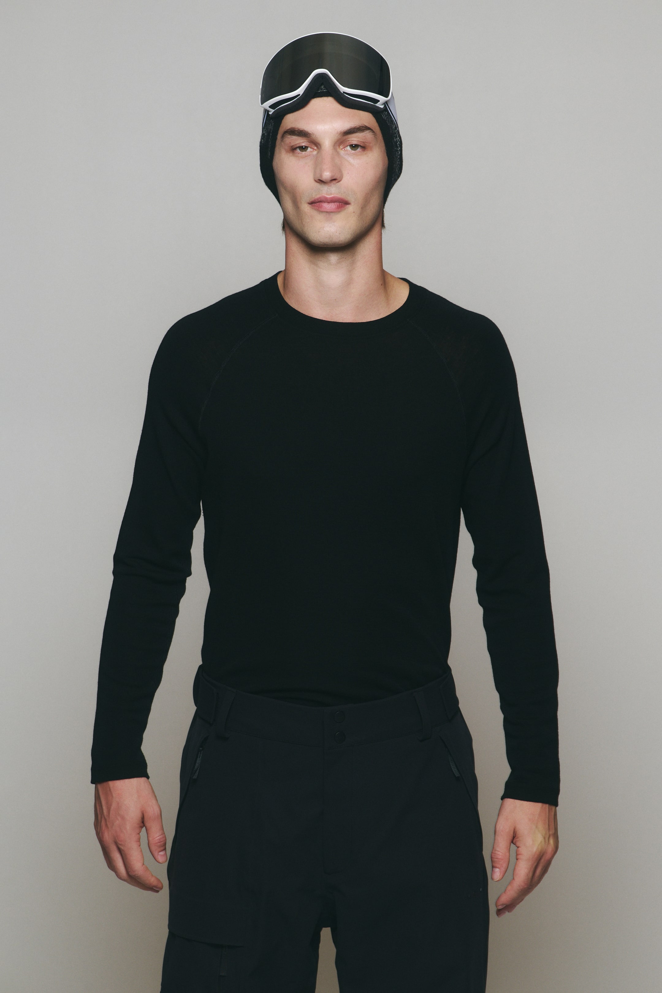 Visualizza l'immagine ingrandita: Un top nero aderente da strato base, con scollo rotondo e maniche lunghe raglan, indossato dalla modella. Il top è infilato in pantaloni da sci neri, completati da un berretto nero e da occhiali da sci con montatura bianca portati sulla testa per un look da sportswear invernale.