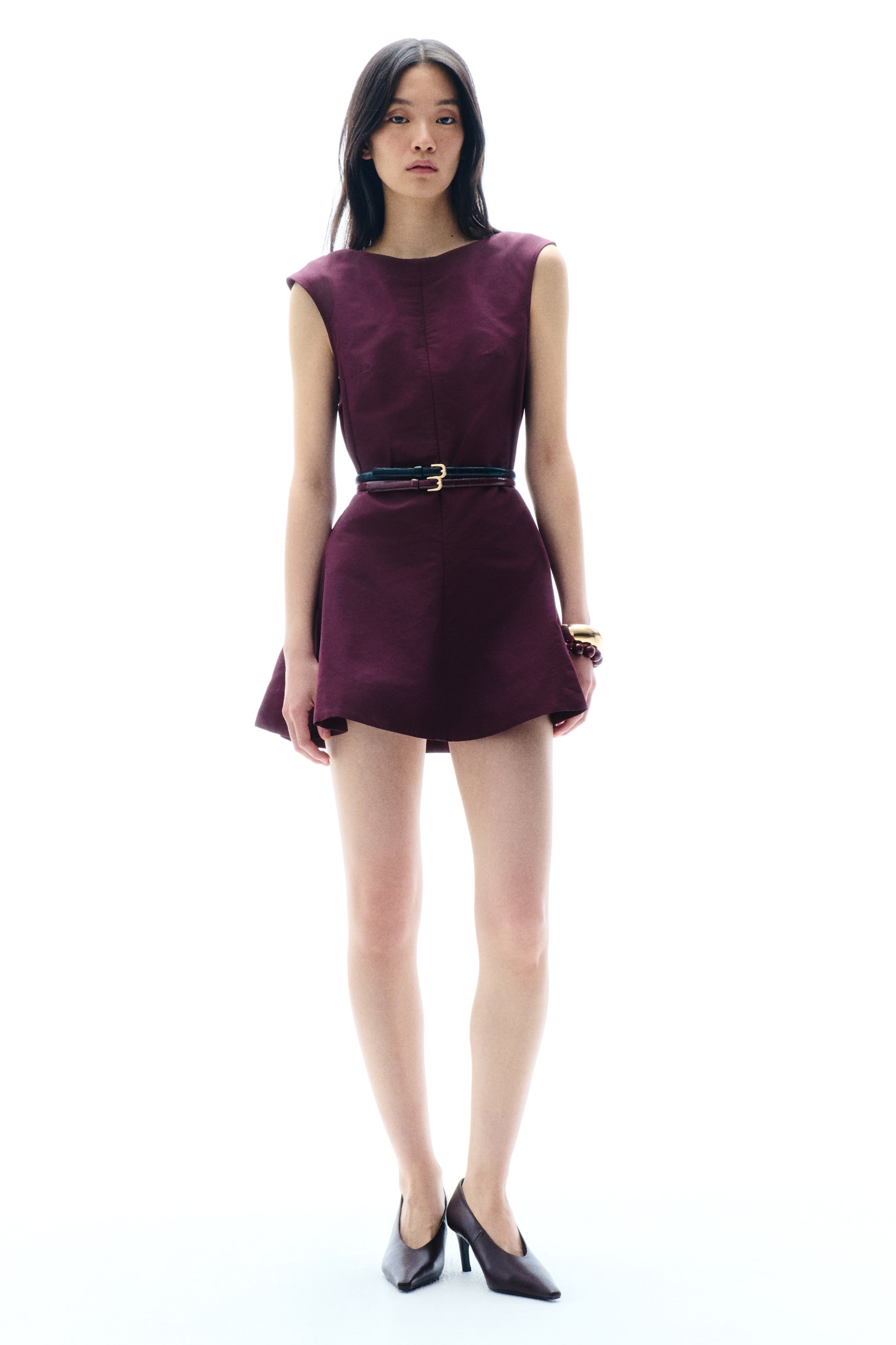 View larger image: A deep plum sleeveless mini dress featuring a round neckline and structured shoulders, cinched at the waist with a slim dark belt with a gold buckle, and flaring out into an A-line skirt.
