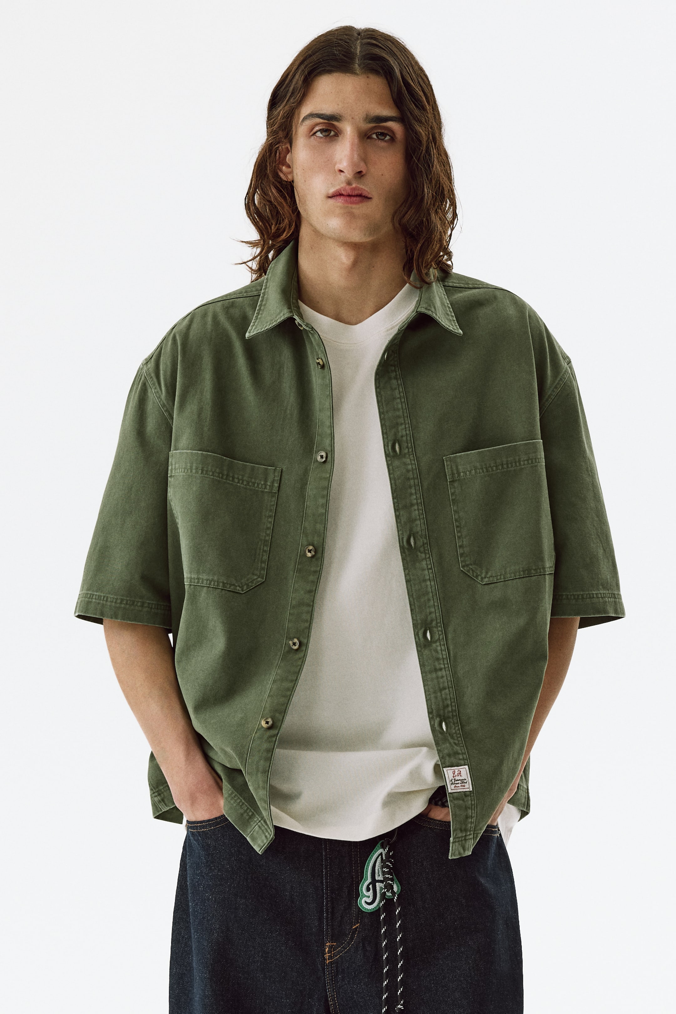View larger image: An olive green short-sleeved shirt, showcasing a relaxed fit, a spread collar, a front button placket with visible buttons, and two patch pockets on the chest, is worn open over a light-coloured t-shirt.