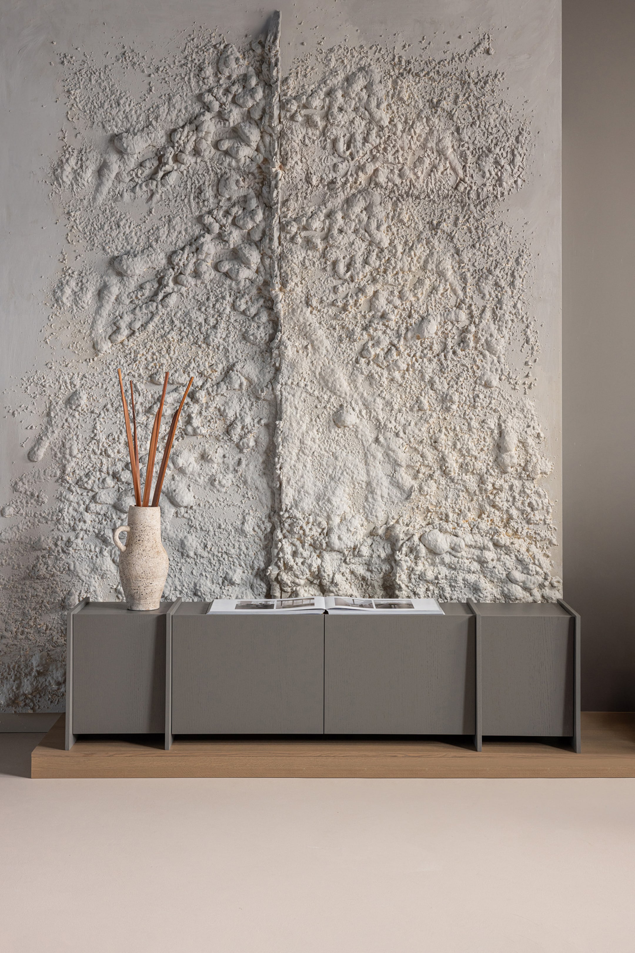 Grotere afbeelding bekijken: Een grijze houten console presenteert een vooraanzicht, rustend op een lichte houten sokkel en heeft een lang, laag ontwerp met twee centrale deurpanelen en stevige eindsecties. Bovenop bevindt zich een gestructureerde vaas met gedroogde rietstengels en een open boek.