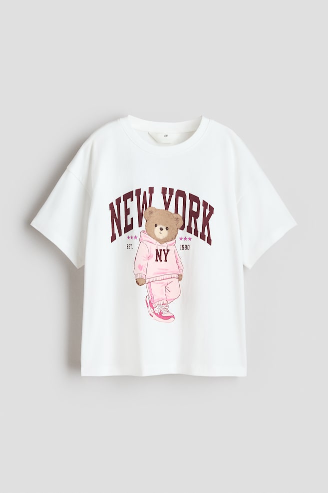 A white T-shirt crafted from printed cotton jersey features a round, rib-trimmed neckline and dropped shoulders. The front showcases a large graphic print of "NEW YORK" in dark red arched lettering above a brown teddy bear dressed in a pink hooded tracksuit and trainers. - 1