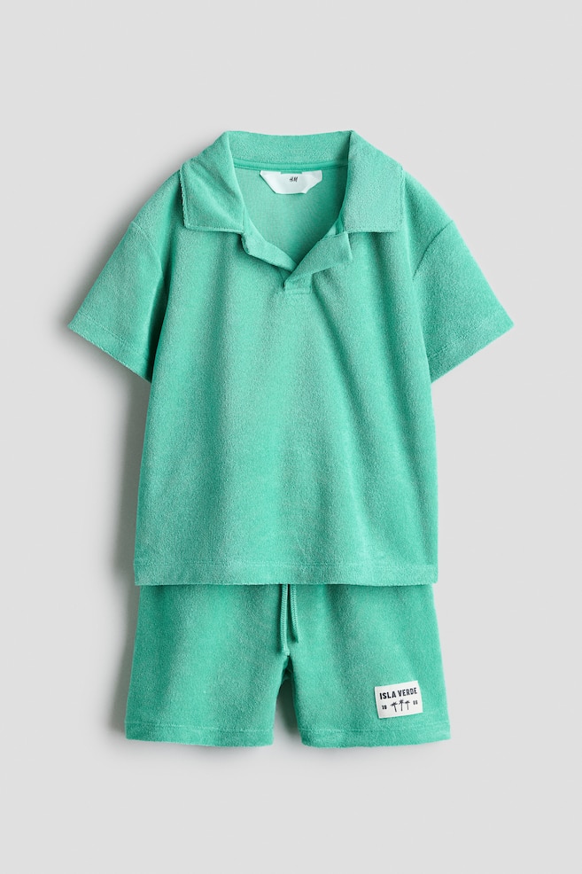 A green two-piece set in soft velour is showcased from the front, featuring a short-sleeved polo shirt with a collar and a V-shaped opening. The coordinating shorts display an elasticated, drawstring waist and a decorative patch on the left leg. - 1