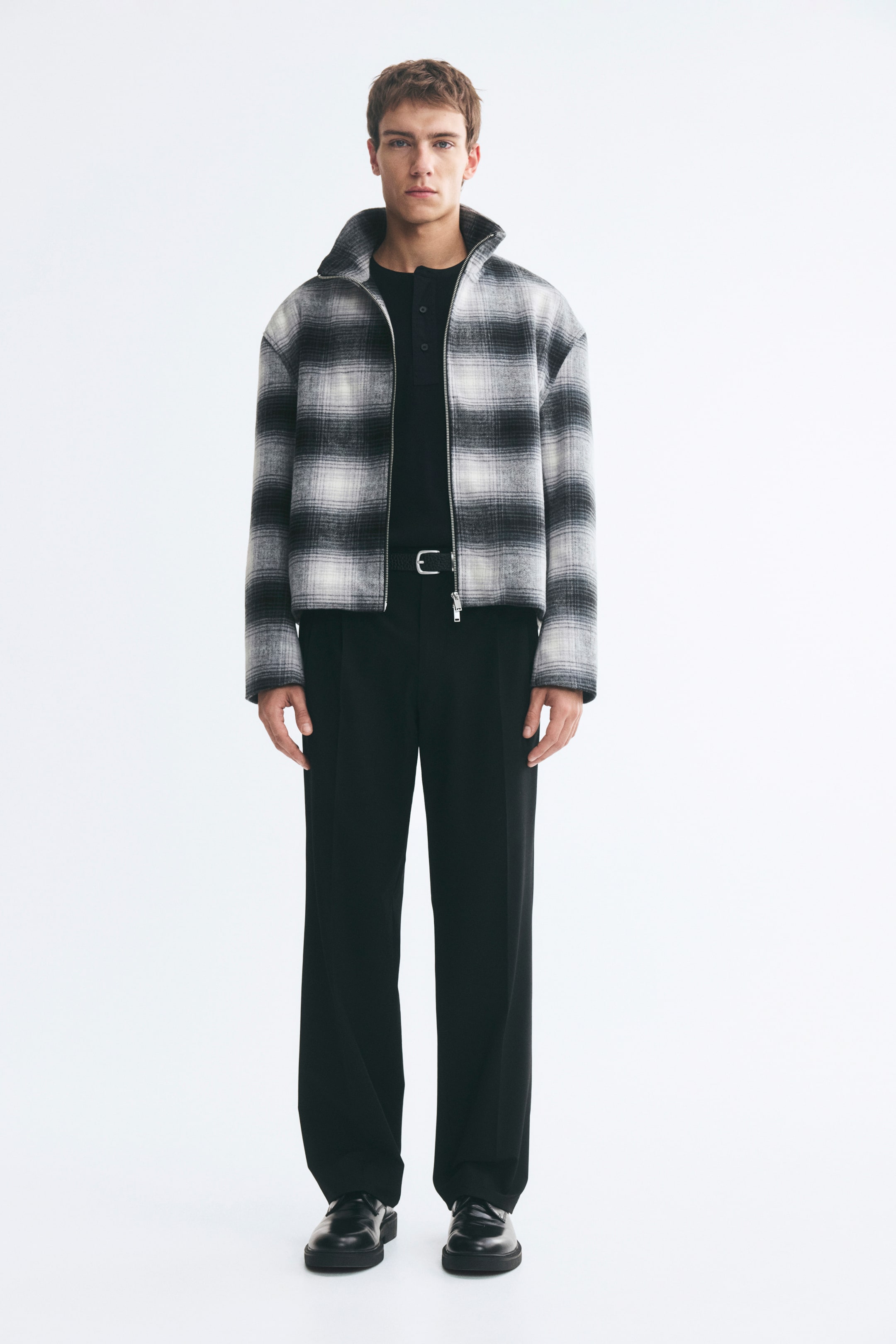 View larger image: A plaid overshirt in black and grey felted fabric is worn by the model, featuring a high stand collar, dropped shoulders, and a silver two-way front zipper left partially open. The overshirt is layered over a black shirt and black trousers.