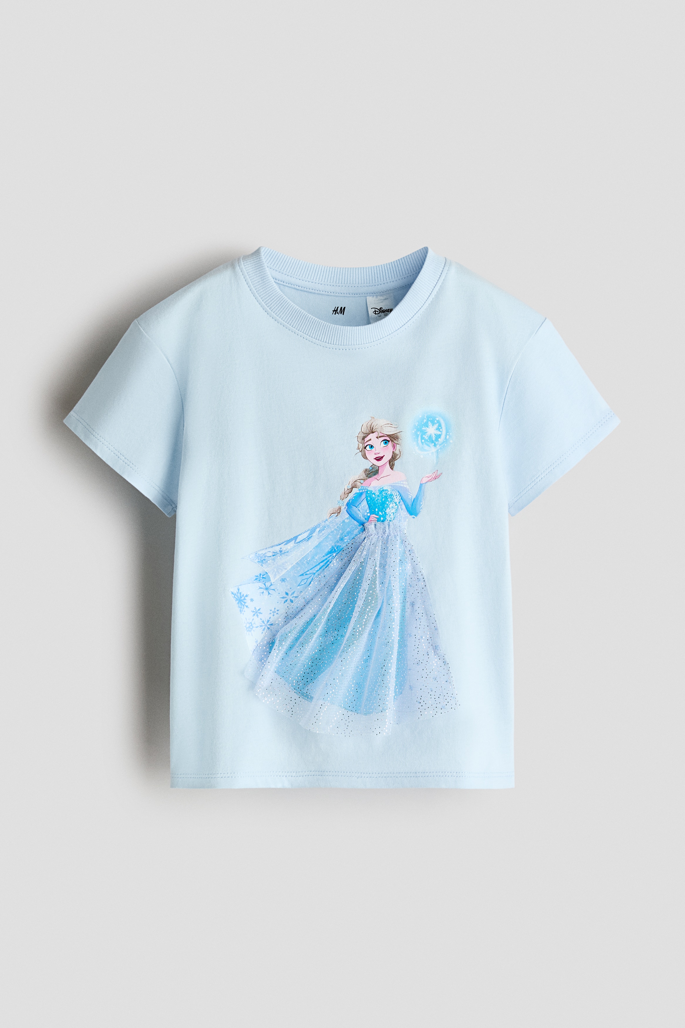 The model is wearing a light blue, short-sleeved T-shirt featuring a character graphic on the front. The motif is embellished with a sheer, glittery blue tulle overlay that forms the characterâs skirt.