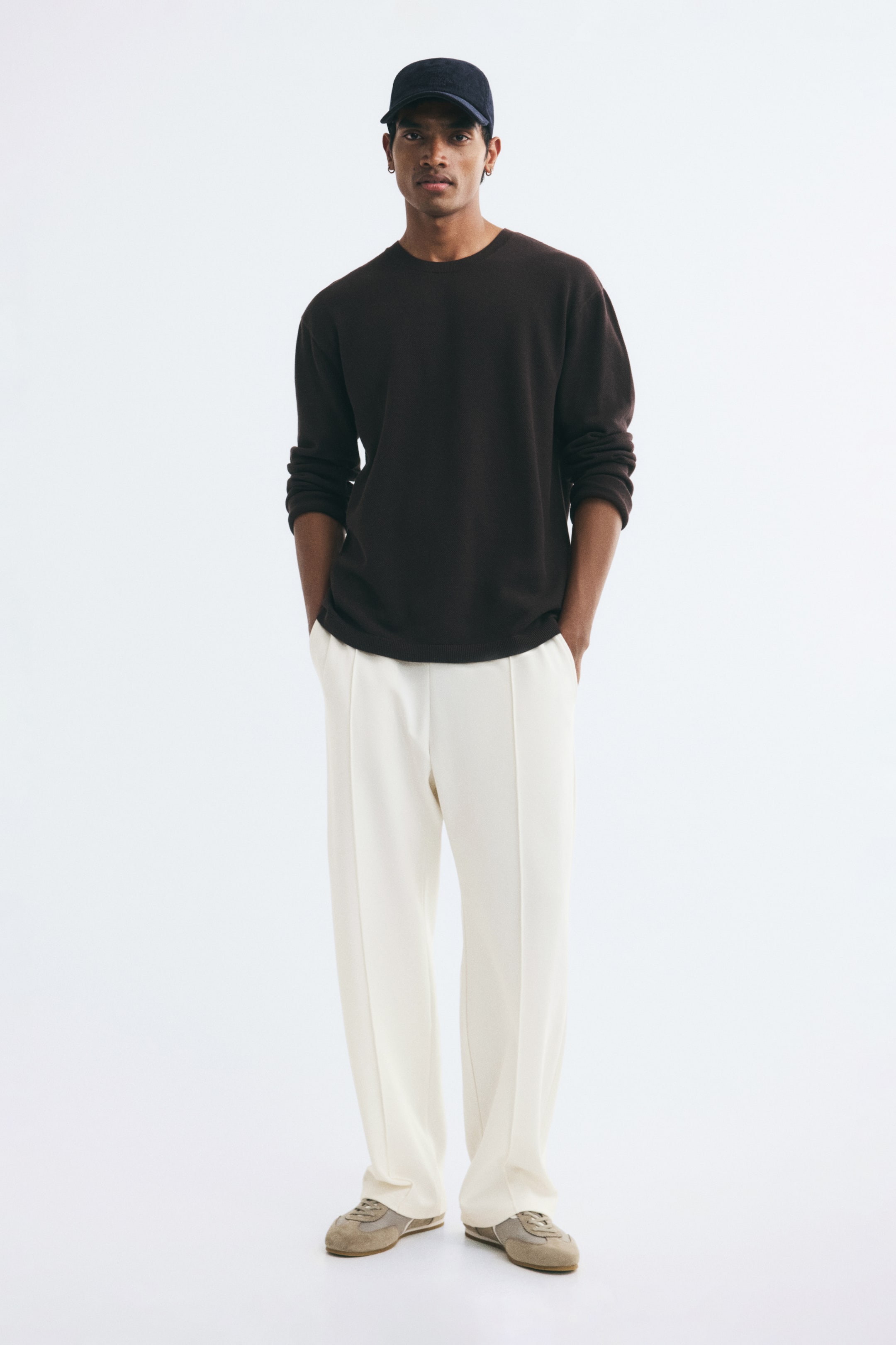View larger image: The model is wearing lightweight jersey joggers in an off-white shade, standing with hands placed in the diagonal side pockets. The relaxed-fit joggers feature straight legs with prominent stitched creases down the front and are styled with a dark brown long-sleeved top and sneakers.