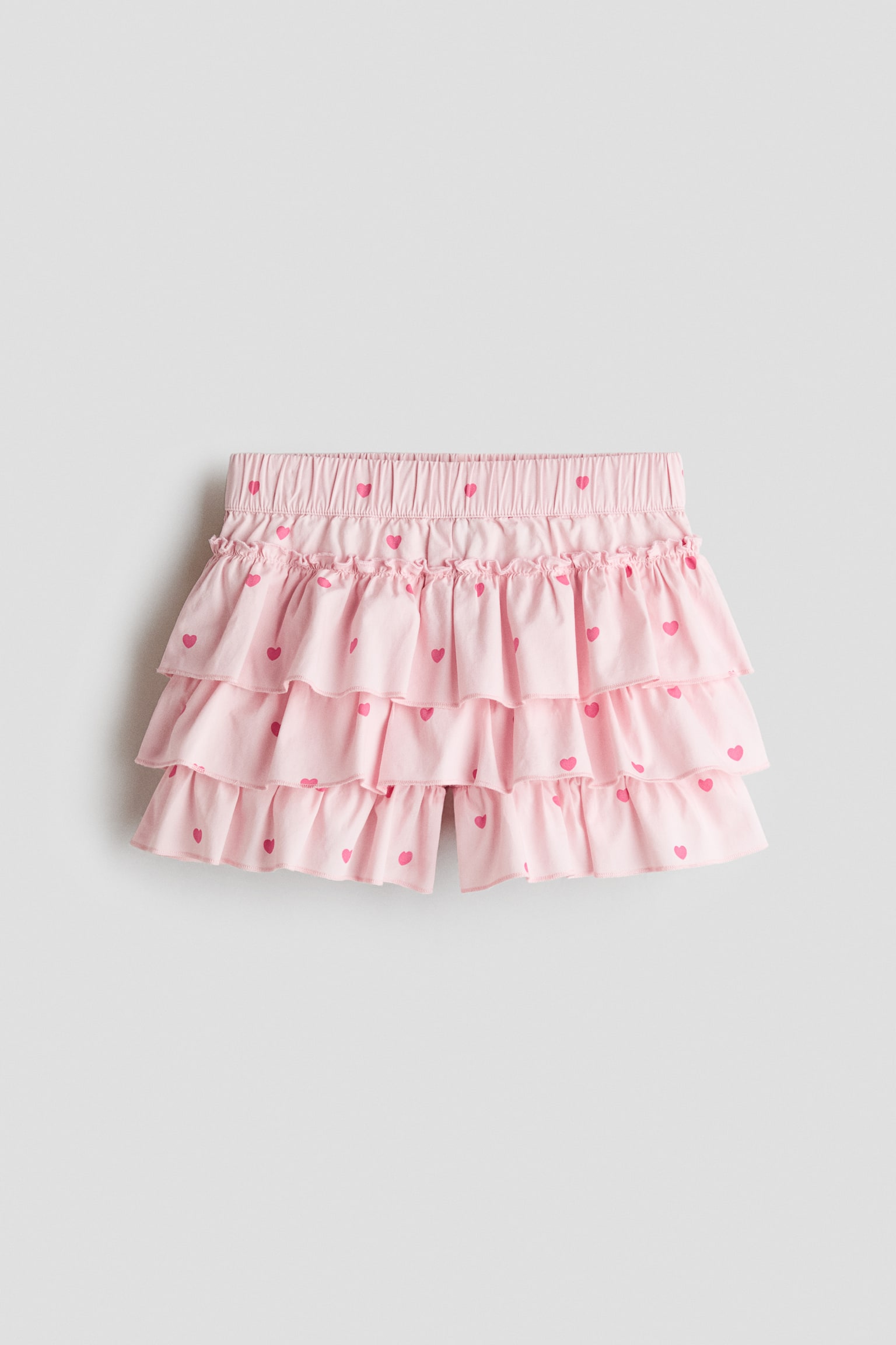 A light pink tiered skort featuring a pattern of small pink hearts is shown from the front, displaying an elasticated waistband with a frill trim at the top. The design showcases three gathered ruffle tiers with shorts visible peeking out from underneath the bottom hem.