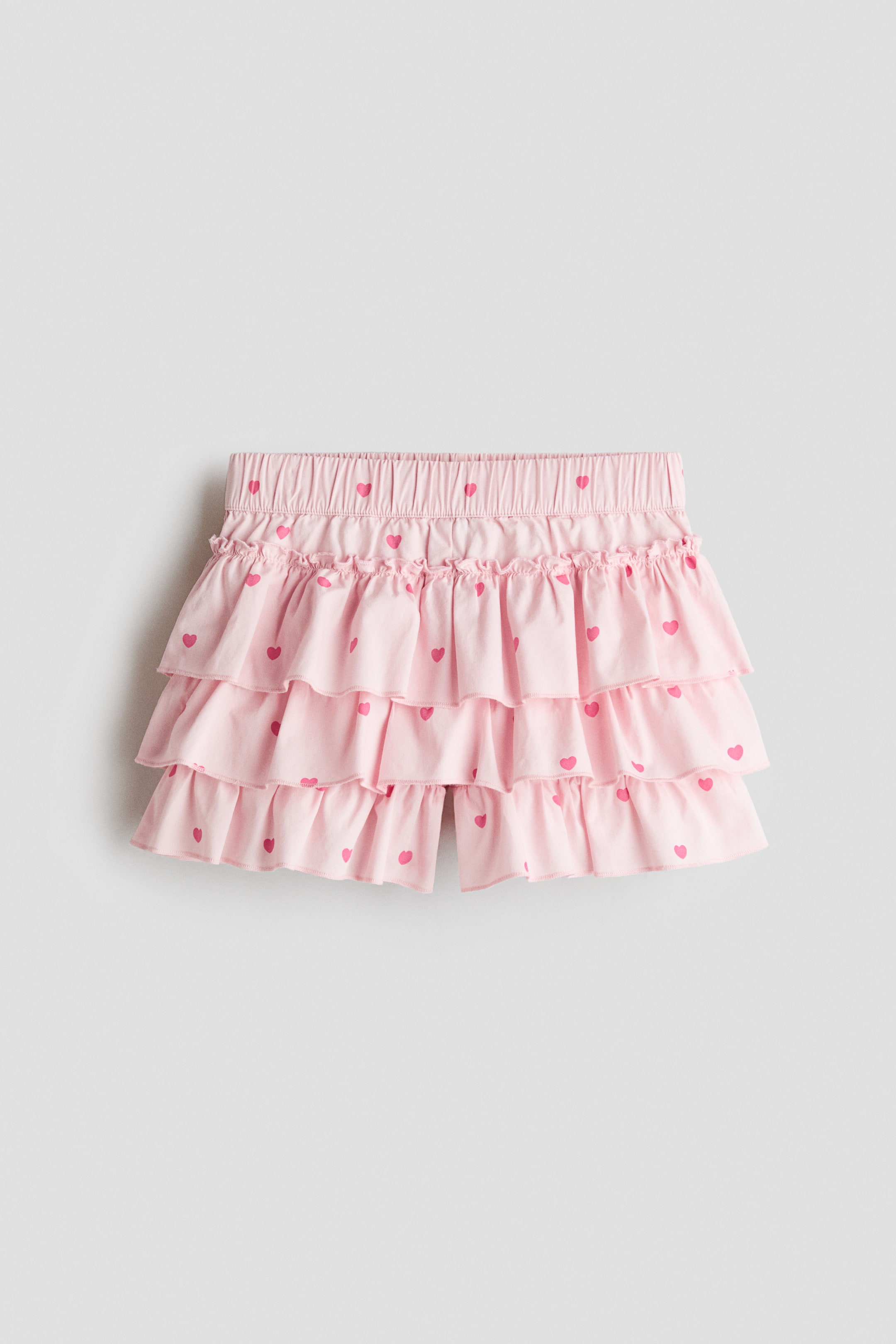 View larger image: A light pink tiered skort featuring a pattern of small pink hearts is shown from the front, displaying an elasticated waistband with a frill trim at the top. The design showcases three gathered ruffle tiers, with shorts visible peeking out from underneath the bottom hem.