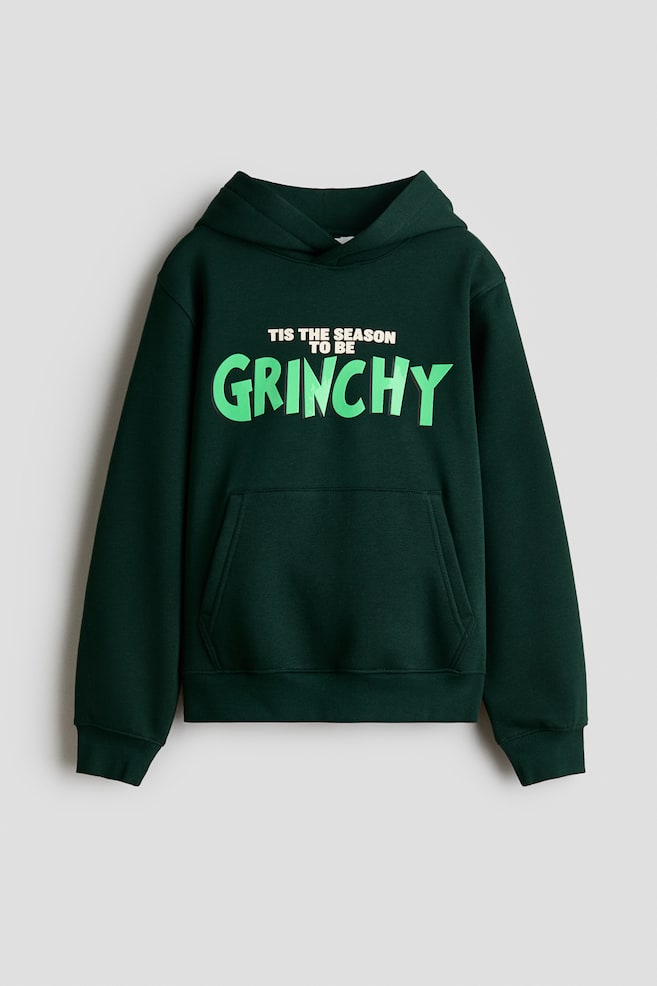 Ein dunkelgrüner Hoodie ist von vorne zu sehen und verfügt über eine Wickelkapuze, überschnittene Schultern, lange Ãrmel und eine Kängurutasche vorne. Auf der Brust ist ein aufgedruckter Schriftzug mit der Aufschrift âTIS THE SEASON TO BE GRINCHYâ in Weià und leuchtendem Grün zu sehen, während gerippte Bündchen und ein gerippter Saum das Design abrunden. - 1