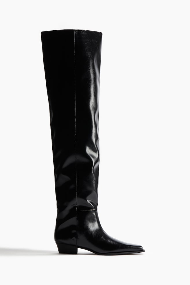 A black, glossy tall boot is standing, featuring a pointed square toe, a low block heel, and a tall shaft that is showing natural creasing. - 1