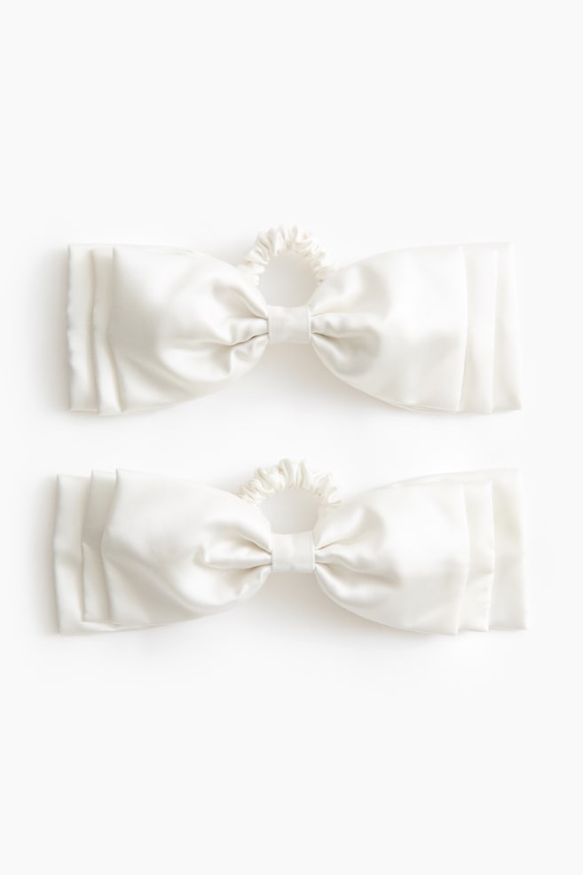 DouÄ accesorii de pÄr tip fundÄ, de culoare alb-deschis Èi cu aspect satinat, sunt aÈezate plat, în vedere frontalÄ, evidenÈiind designul lor cu fundÄ Ã®n straturi multiple. Fiecare accesoriu este realizat dintr-un material neted, lucios, cu aripi structurate, stratificate, pe ambele pÄrÈi ale unui nod central Èi cu o micÄ bandÄ elasticÄ plisatÄ ataÈatÄ Ã®n partea superioarÄ. - 1