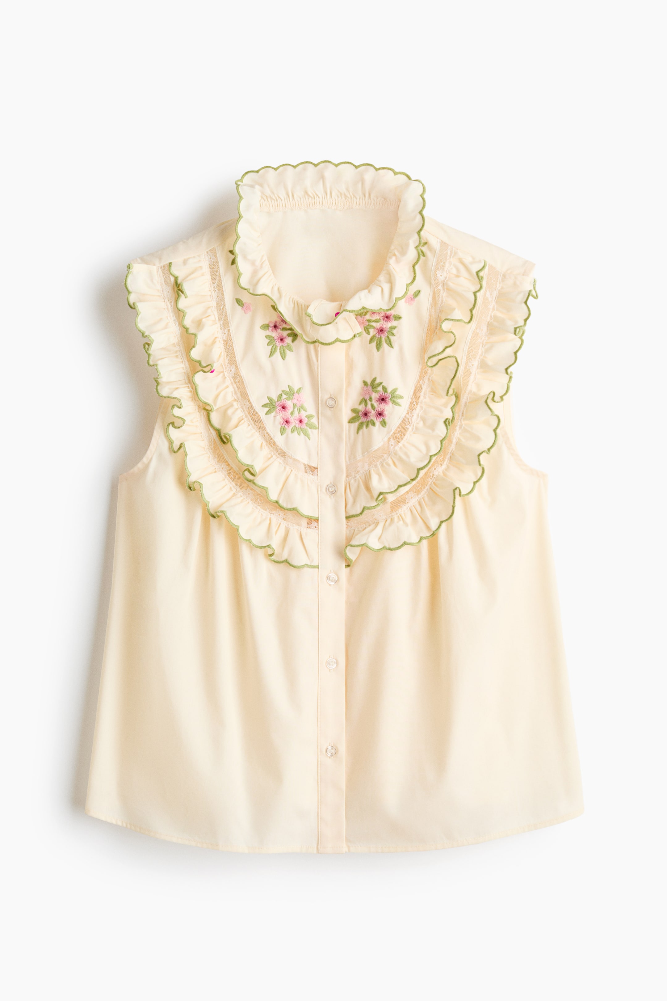 Xem ảnh lớn hơn: Áo blouse không tay màu vàng nhạt có cổ cao bèo nhún và hai tầng bèo viền xanh lá cây với chi tiết ren trắng chạy dọc phía trước, kéo dài qua vai. Thêu hoa màu hồng và xanh lá cây trang trí khu vực ngực giữa, phía trên hàng nút phía trước.
