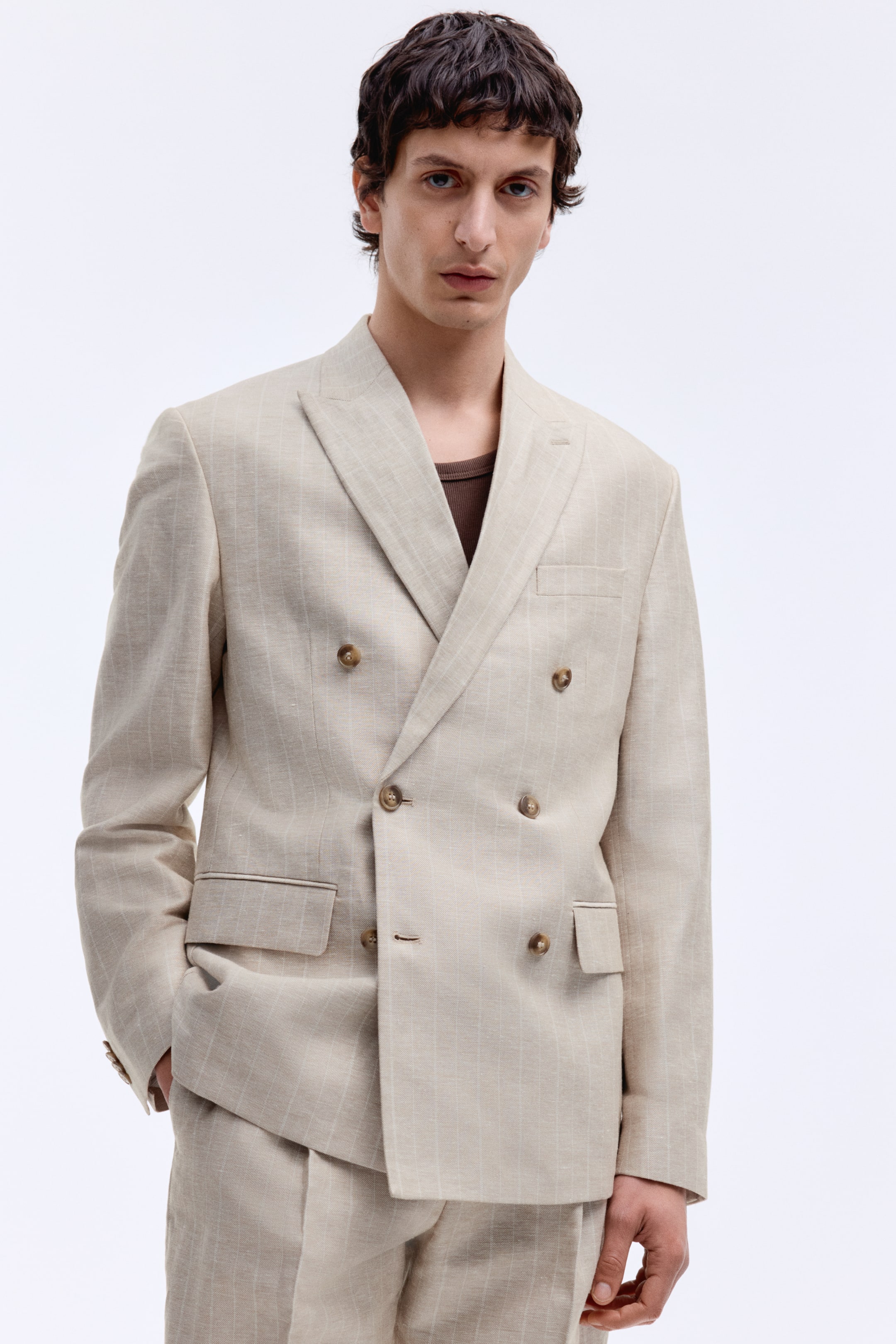 View larger image: The model is wearing a double-breasted jacket in a light beige pinstriped weave, featuring peak lapels, a chest pocket, two flap front pockets, and decorative cuff buttons. The model's hand is placed in a front pocket, with the jacket styled over a brown top and paired with matching trousers.