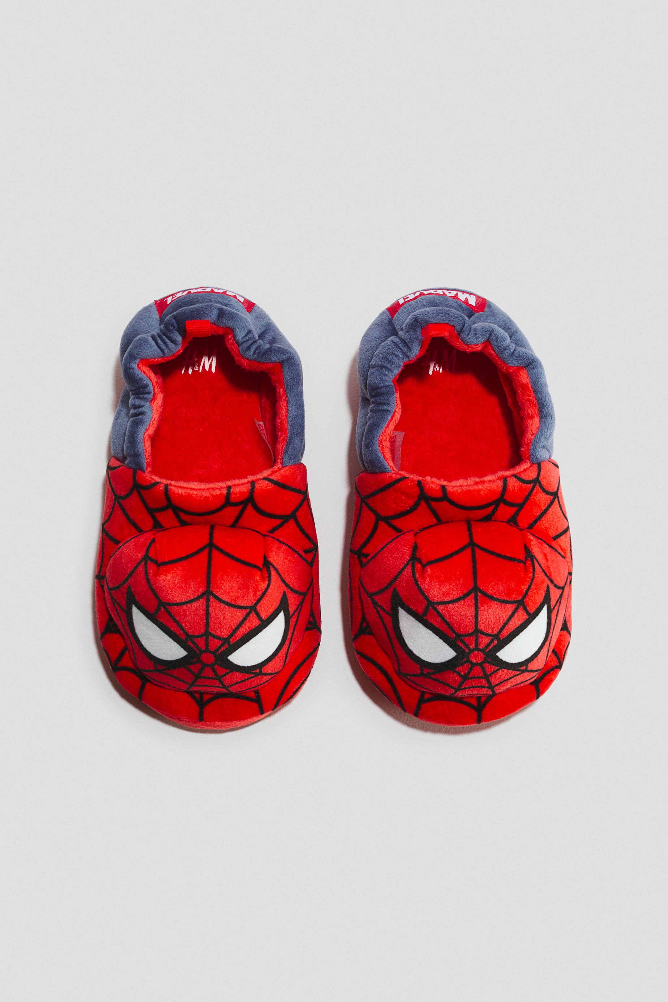 Zobraziť väčší obrázok: Pár červených a modrých papúč Spider-Man je umiestnený vedľa seba, smerujúc dopredu. Každá papuča má červený plyšový vrch s čiernym vzorom pavučiny a bielymi očami Spider-Mana, modrý elastický golier na členku odhaľujúci červenú plyšovú podšívku a viditeľné značky 'H&M' a 'Marvel' na vnútornej strane a vonkajšom zadnom okraji.