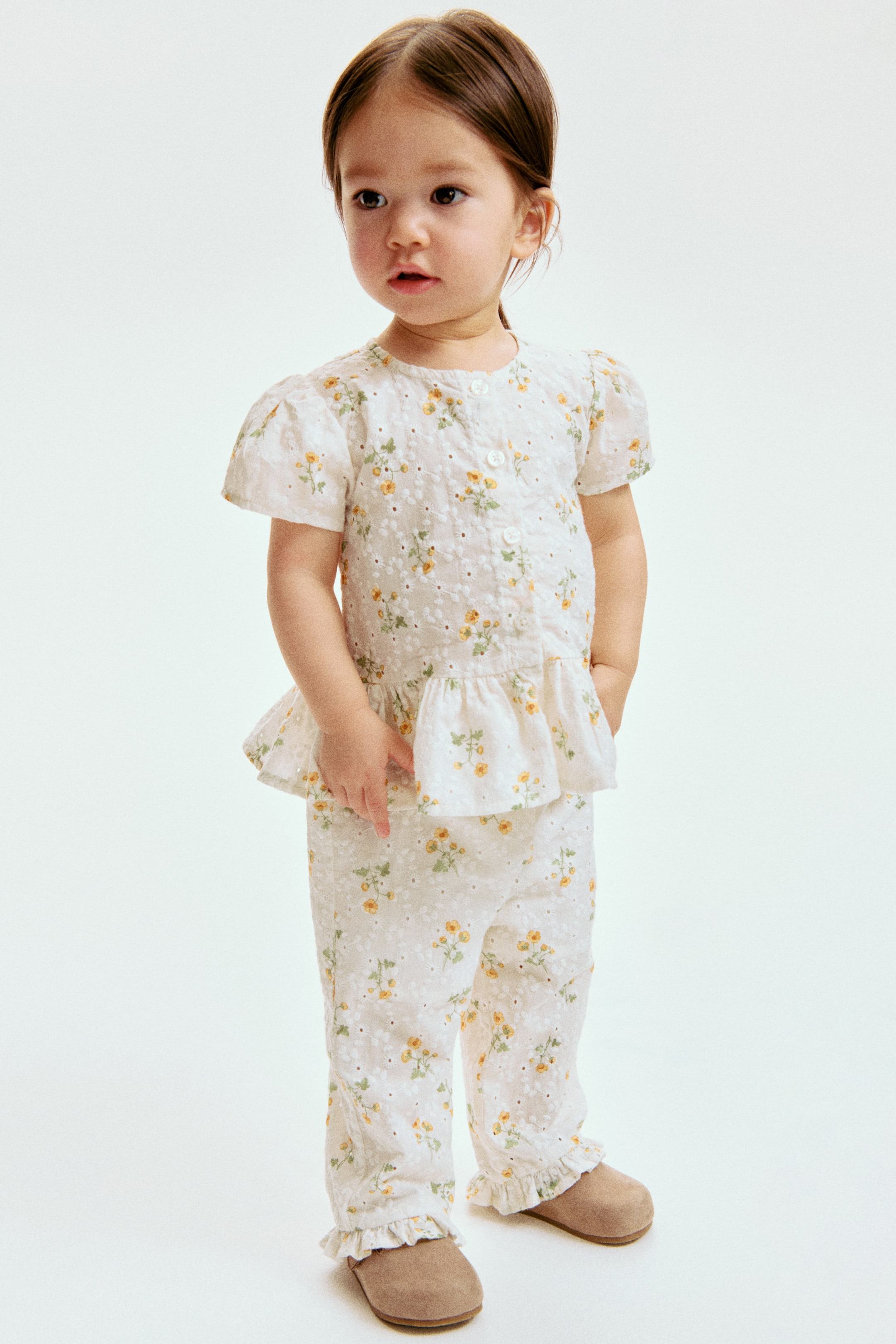 Ampliar la imagen: Un conjunto de dos piezas en blanco presenta un patrón de bordado floral amarillo y de ojales. La blusa de manga corta está diseñada con mangas abullonadas, un escote redondeado con cierre de botones y un dobladillo de peplum con volantes, mientras que los pantalones a juego muestran puños con volantes en los tobillos.