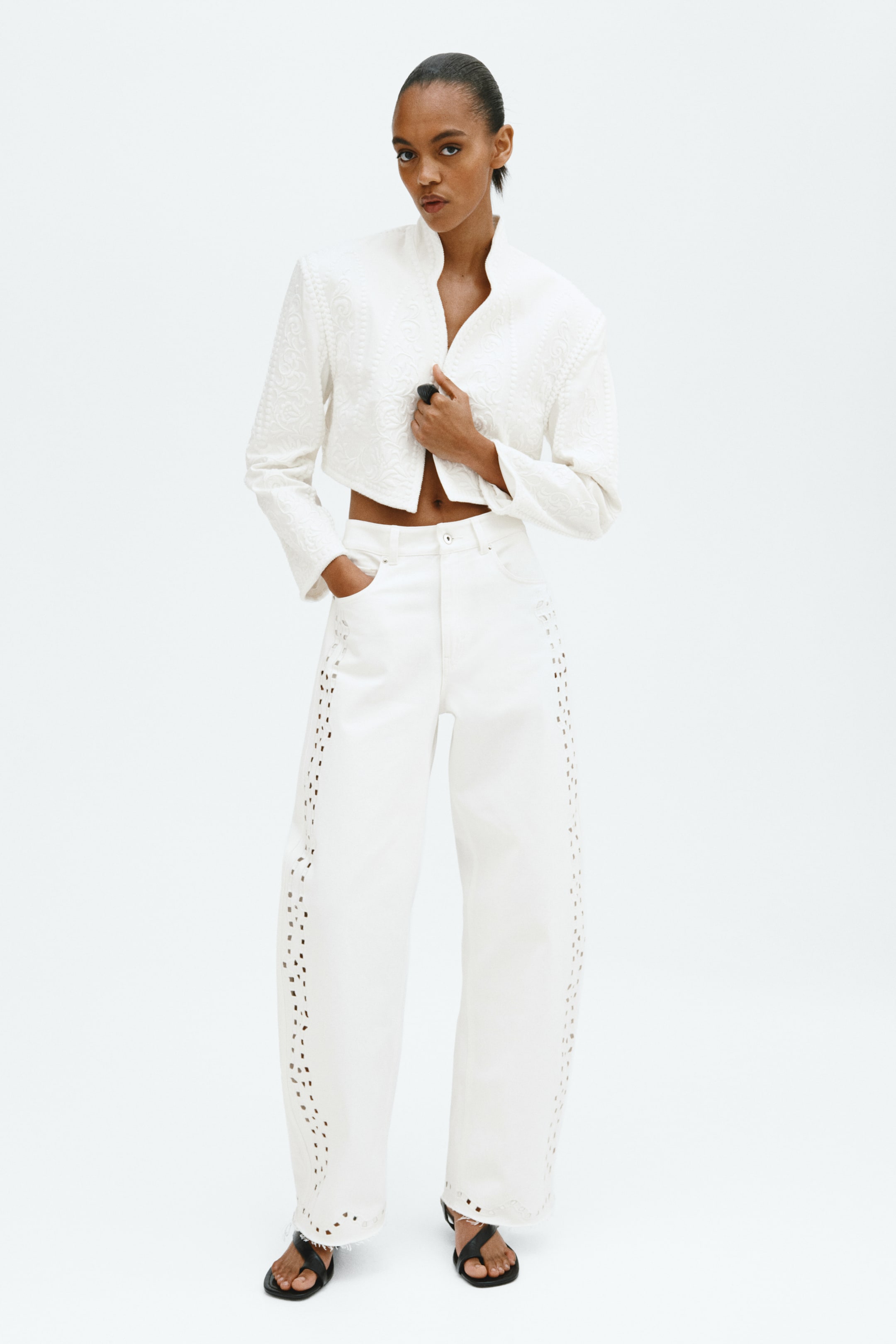 View larger image: Presenting a white cropped jacket with an embossed floral pattern, structured shoulders, and a standing collar. It's paired with white wide-leg pants featuring laser-cut detailing along the side seams and a frayed hem.