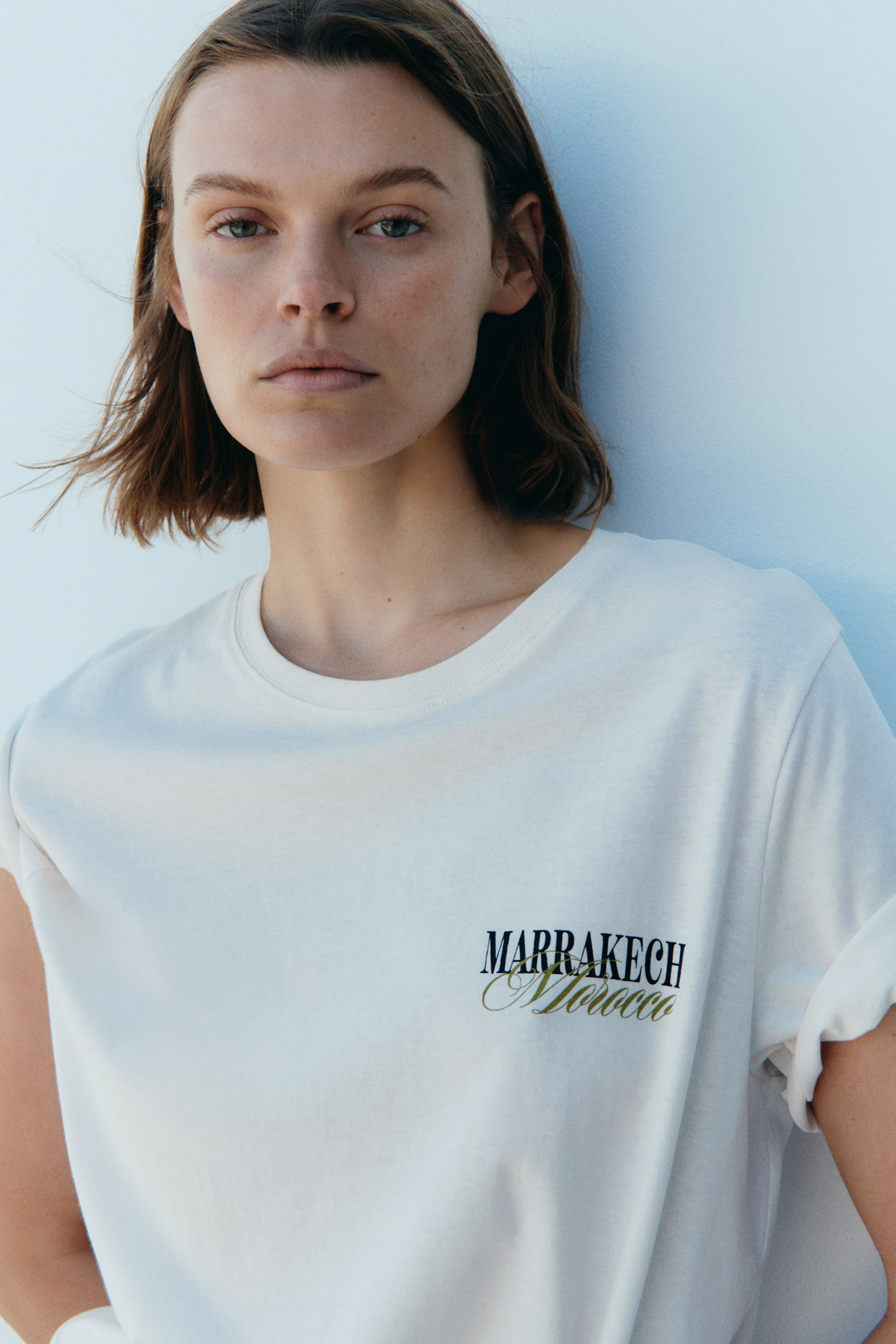 View larger image: The model is dressed in an off-white classic t-shirt featuring a round neckline and printed motifs. The motifs showcase "MARRAKECH" in bold black lettering and "Morocco" in elegant gold script across the chest, with the model opting for cuffed short sleeves.