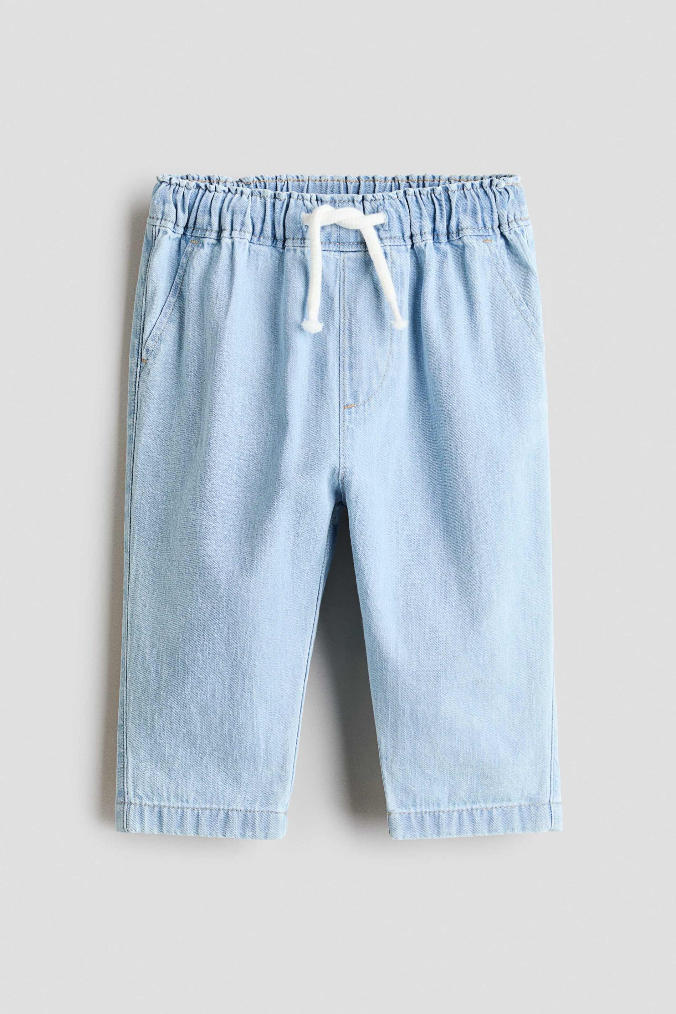 Größeres Bild ansehen: Ein Paar hellblaue Denim-Hosen ist von vorne zu sehen, mit einem elastischen, gerafften Bund, der mit einem weißen Kordelzug in der Mitte gebunden ist. Zwei vordere Schlitztaschen sind sichtbar, und die Hosen sind mit umgeschlagenen Säumen abgeschlossen.