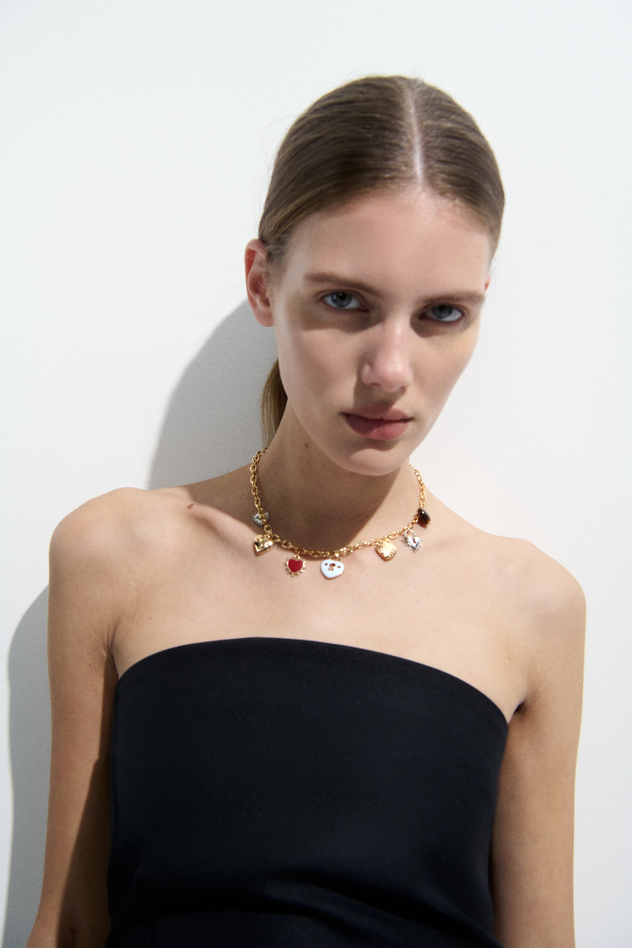 View larger image: A gold-toned chain necklace features an assortment of charms, including red and white enameled hearts, a silver broken heart, a gold shell, a dark brown oval, and a winged gold heart. The necklace rests at the base of the neck, displaying the charms along its length.