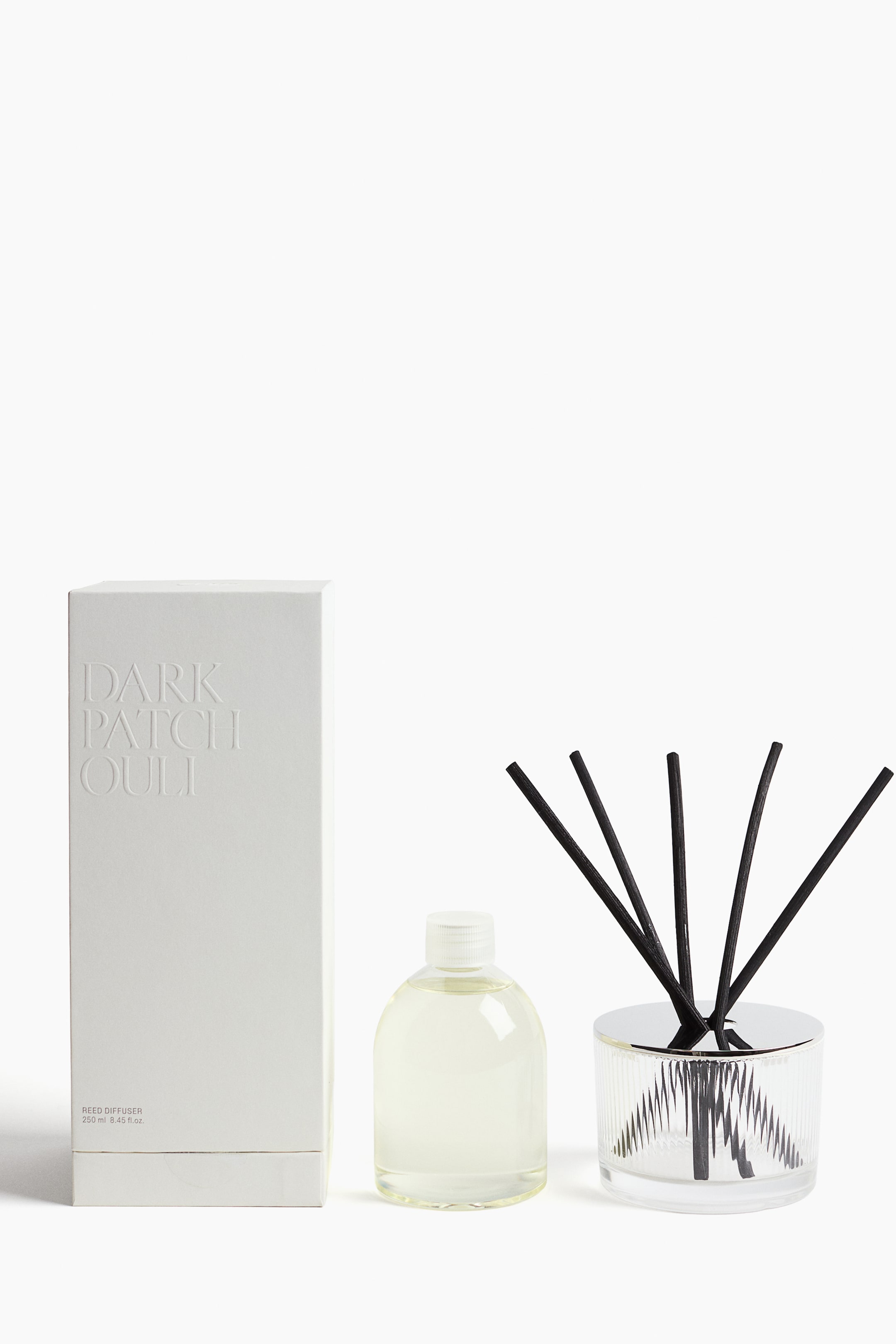 Visualizza l'immagine ingrandita: Un set di diffusori a bastoncini è esposto frontalmente, composto da una scatola bianca testurizzata con la scritta in rilievo “DARK PATCHOULI” a sinistra, un flacone di ricarica in plastica trasparente contenente un liquido giallo pallido al centro e un recipiente in vetro trasparente zigrinato con coperchio lucido color argento che contiene cinque bastoncini diffusori neri a destra.