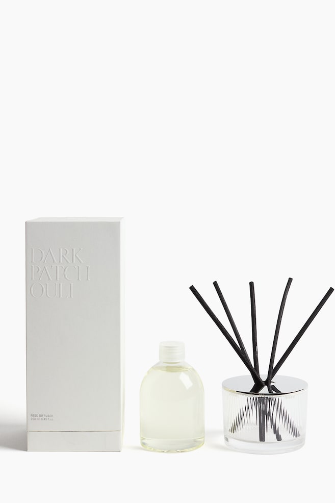 A reed diffuser set is displayed facing front, consisting of a white textured box with embossed "DARK PATCHOULI" text on the left, a clear plastic refill bottle containing pale yellow liquid in the center, and a ribbed clear glass vessel with a polished silver-toned lid holding five black diffuser reeds on the right. - 1