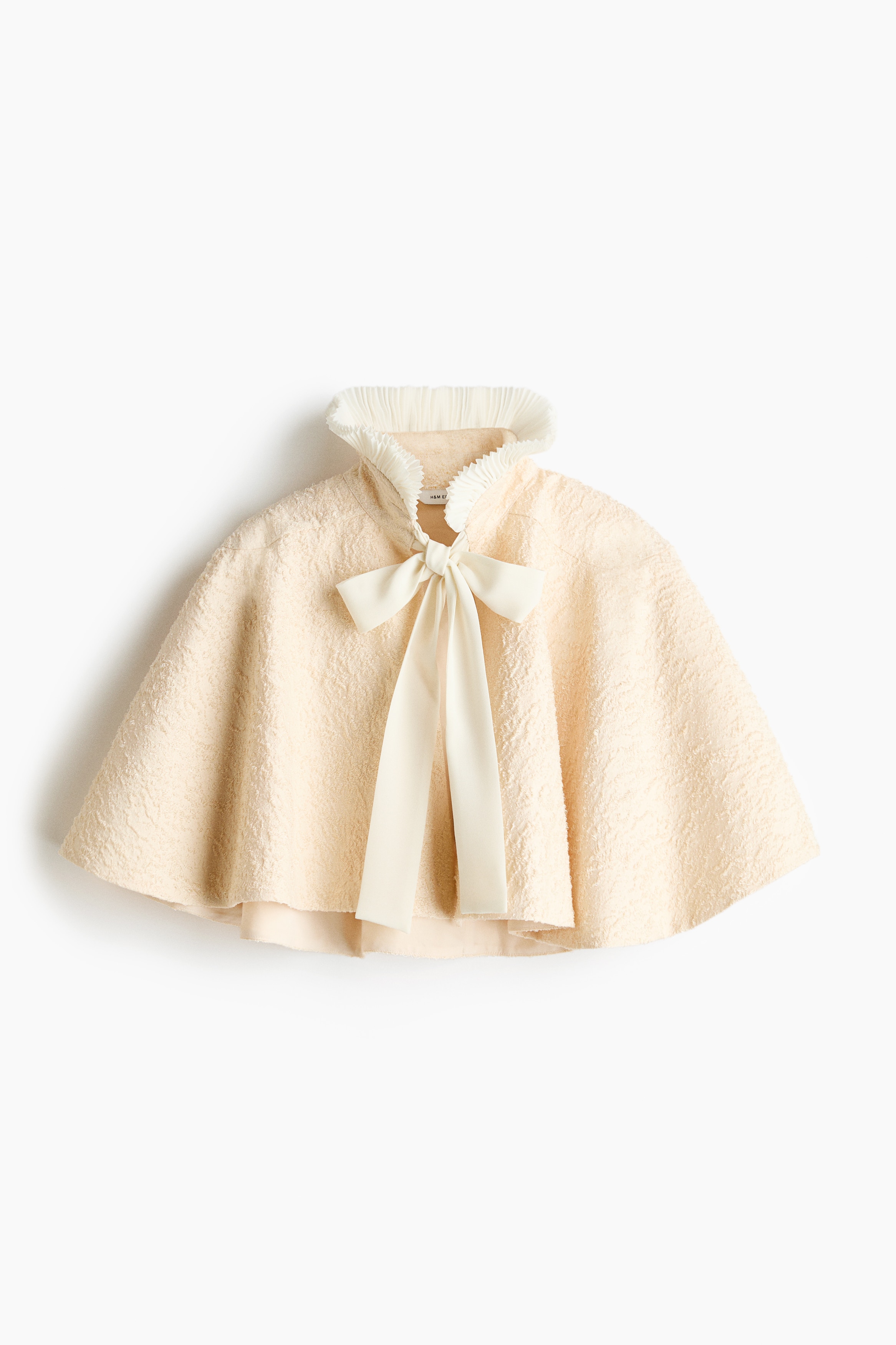 Une cape courte texturÃ©e couleur crÃ¨me claire, dotÃ©e dâun col montant Ã  volants et dâune fermeture par liens sur le devant, est portÃ©e par le mannequin. La cape courte est superposÃ©e Ã  une blouse blanche Ã  manches longues, avec poignets et bas de vÃªtement Ã  volants.