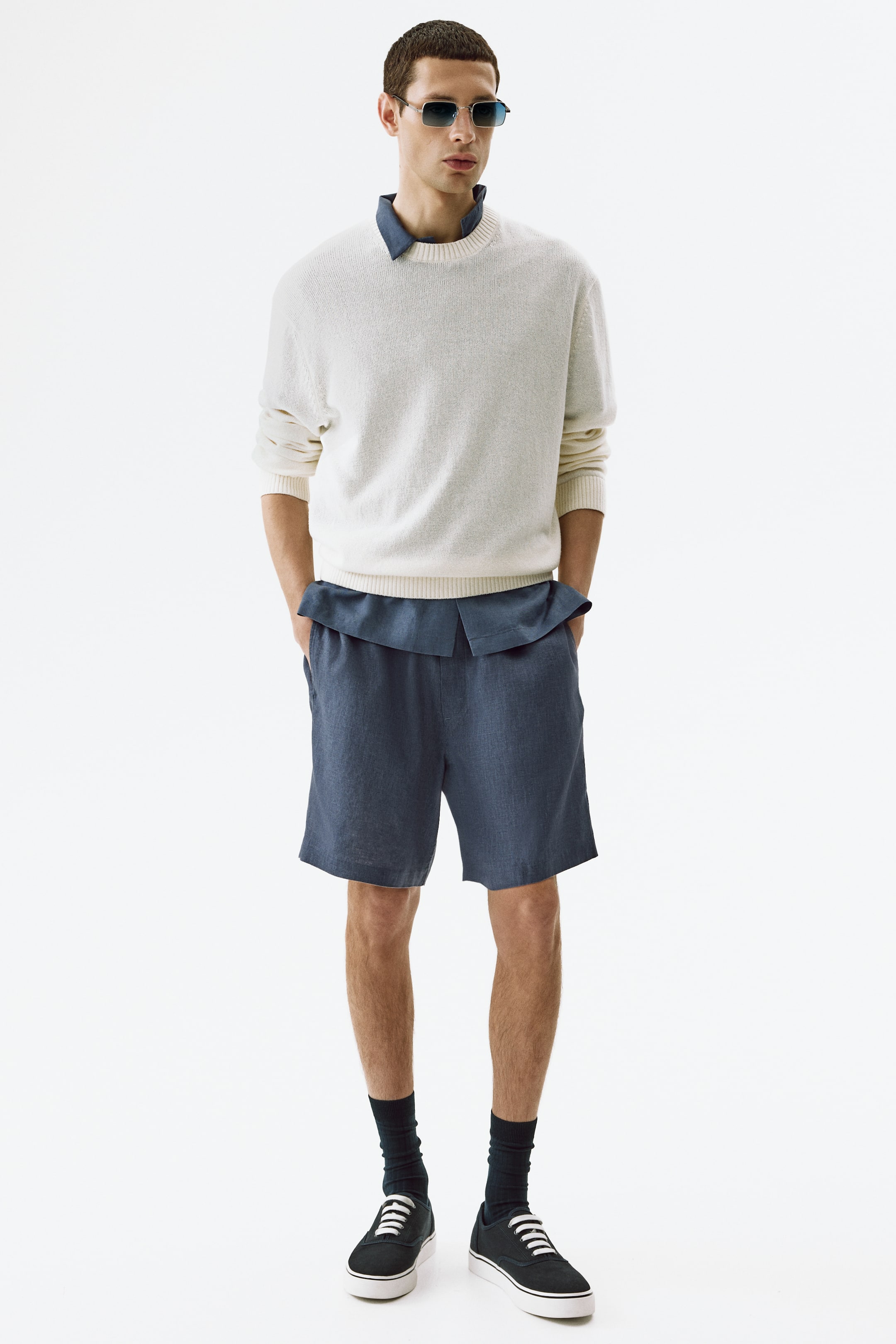 View larger image: The complete outfit is showcased from a front view, featuring an off-white textured crew-neck sweater with ribbed cuffs and hem, layered over a dark blue collared shirt. Dark blue shorts with an elasticized waistband and side pockets are visible below, styled with dark blue ankle socks and black canvas sneakers with white laces. Square-framed sunglasses with blue tinted lenses rest on the face.