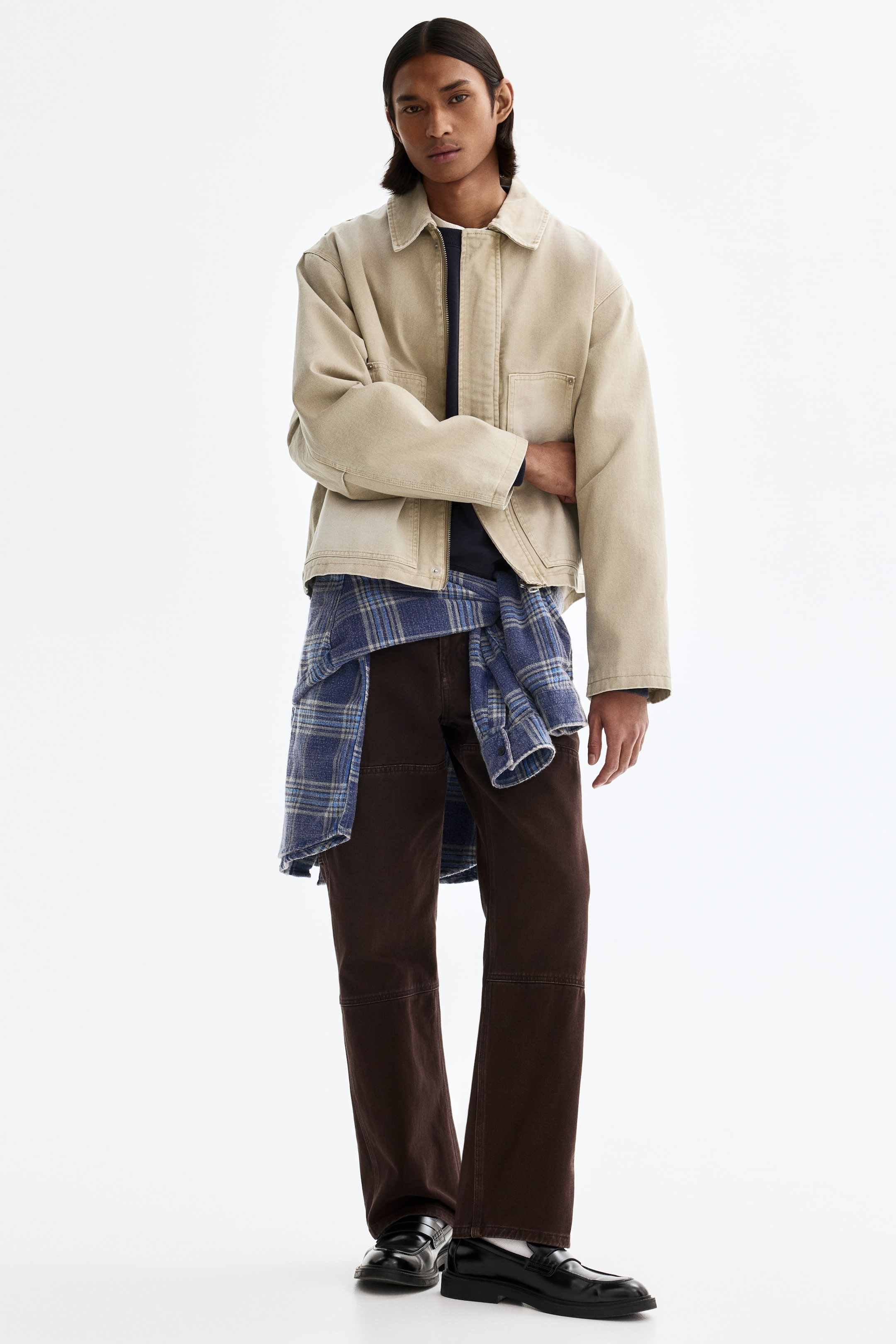 拡大画像を表示: 正面を向いたコーディネートは、フロントがジッパー仕様でクラシックカラーのベージュのオーバーサイズワーカージャケットを主役に、ダークブルーのトップスを重ね、ブラウンのパネルパンツ、ウエストに巻いたブルーのチェックフランネルシャツ、黒のローファーを合わせています。