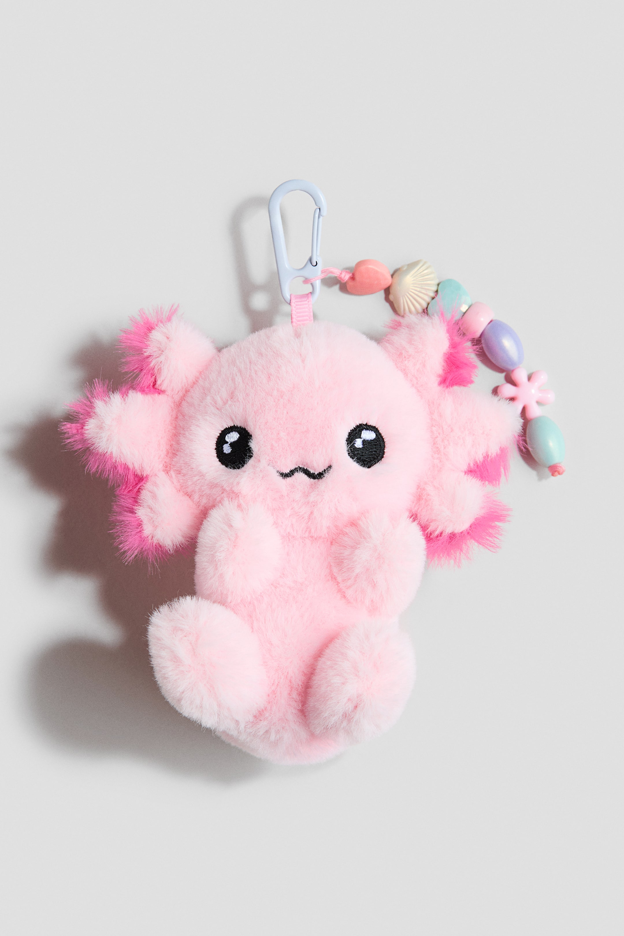 Ver imagen más grande: Un colgante para bolso que presenta un gran juguete de peluche rosa acolchado y suave, mirando al frente, con ojos negros, una boca cosida y estructuras esponjosas similares a branquias en su cabeza. Un mosquetón de metal azul claro está unido en la parte superior, junto con una cadena de cuentas coloridas y con formas.