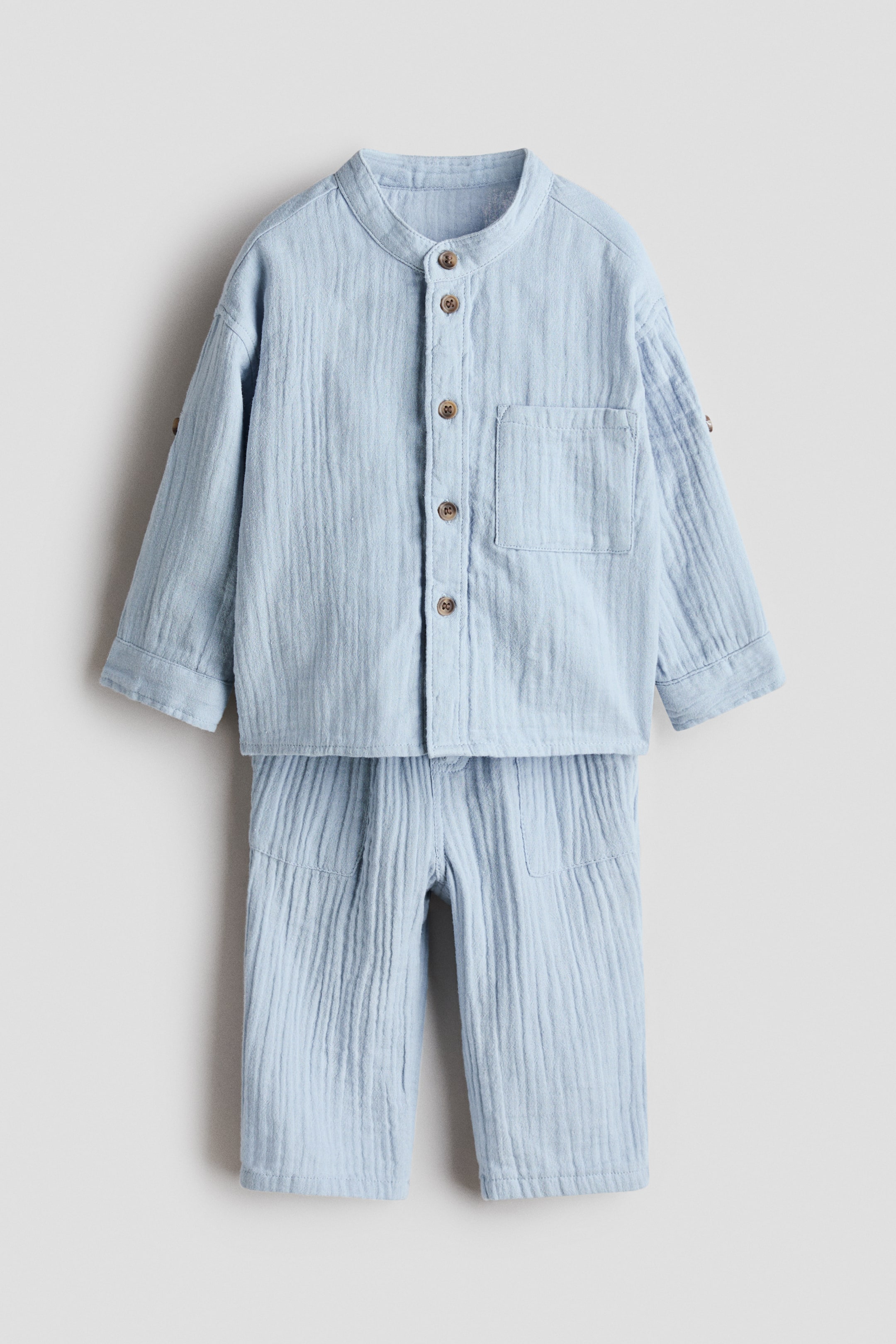 View larger image: A light blue two-piece set, featuring a shirt and trousers, is displayed facing front, all crafted from soft, textured muslin. The shirt has a band collar, buttons running down the front, an open chest pocket, and long sleeves showing tabs with buttons and buttoned cuffs, while the accompanying trousers feature a drawstring waist and side pockets.