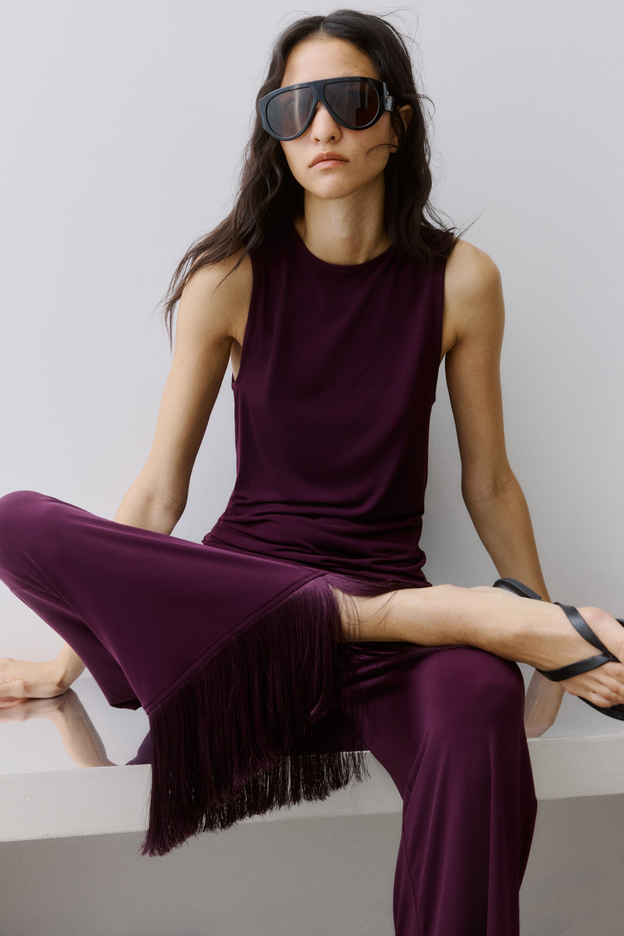 Visualizza immagine più grande: Un top senza maniche color prugna scuro scivola su pantaloni a gamba larga, caratterizzati da lunghe frange coordinate lungo le cuciture esterne delle gambe.