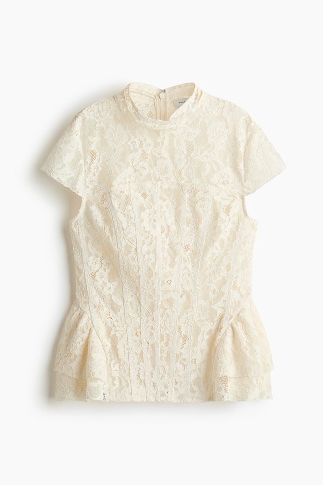 A cream lace top is displayed front-facing, featuring a high neckline, short cap sleeves, and a fitted bodice with vertical seams. It showcases a ruffled peplum hem and a zip fastening visible at the back of the neck, all crafted from floral lace and appears to be lined. - 1