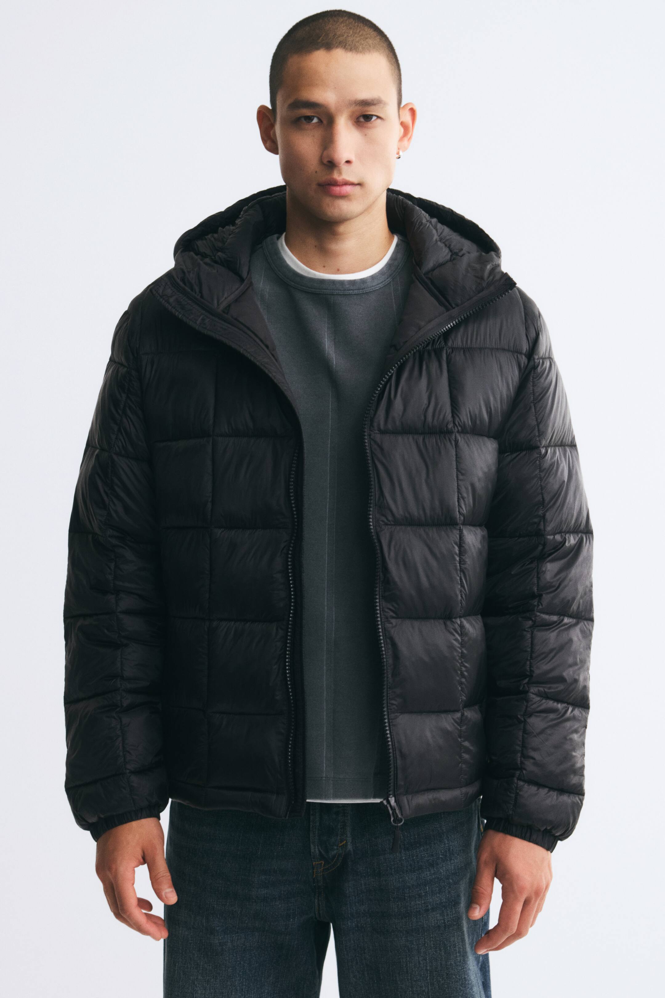 View larger image: A black lightweight puffer jacket featuring a square-quilted pattern is being worn open by the model. The hooded jacket is layered over a grey crew-neck sweatshirt and dark blue jeans.