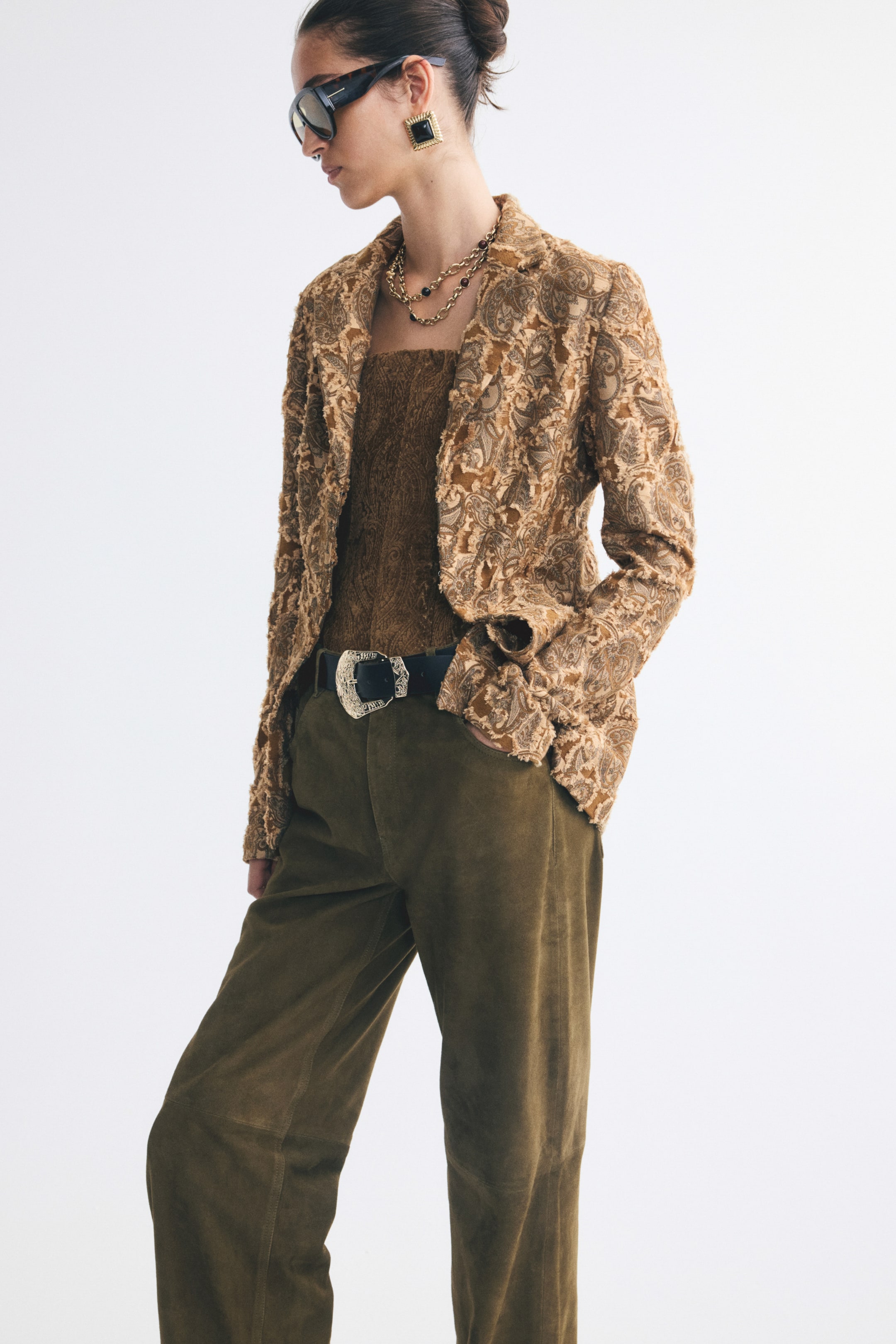 Xem ảnh lớn hơn: Một chiếc blazer một hàng khuy bằng vải jacquard dệt nổi màu nâu vàng với họa tiết paisley và ve áo notch đang được người mẫu mặc mở. Blazer được phối cùng áo ống đồng bộ bên trong và quần màu xanh ô liu.