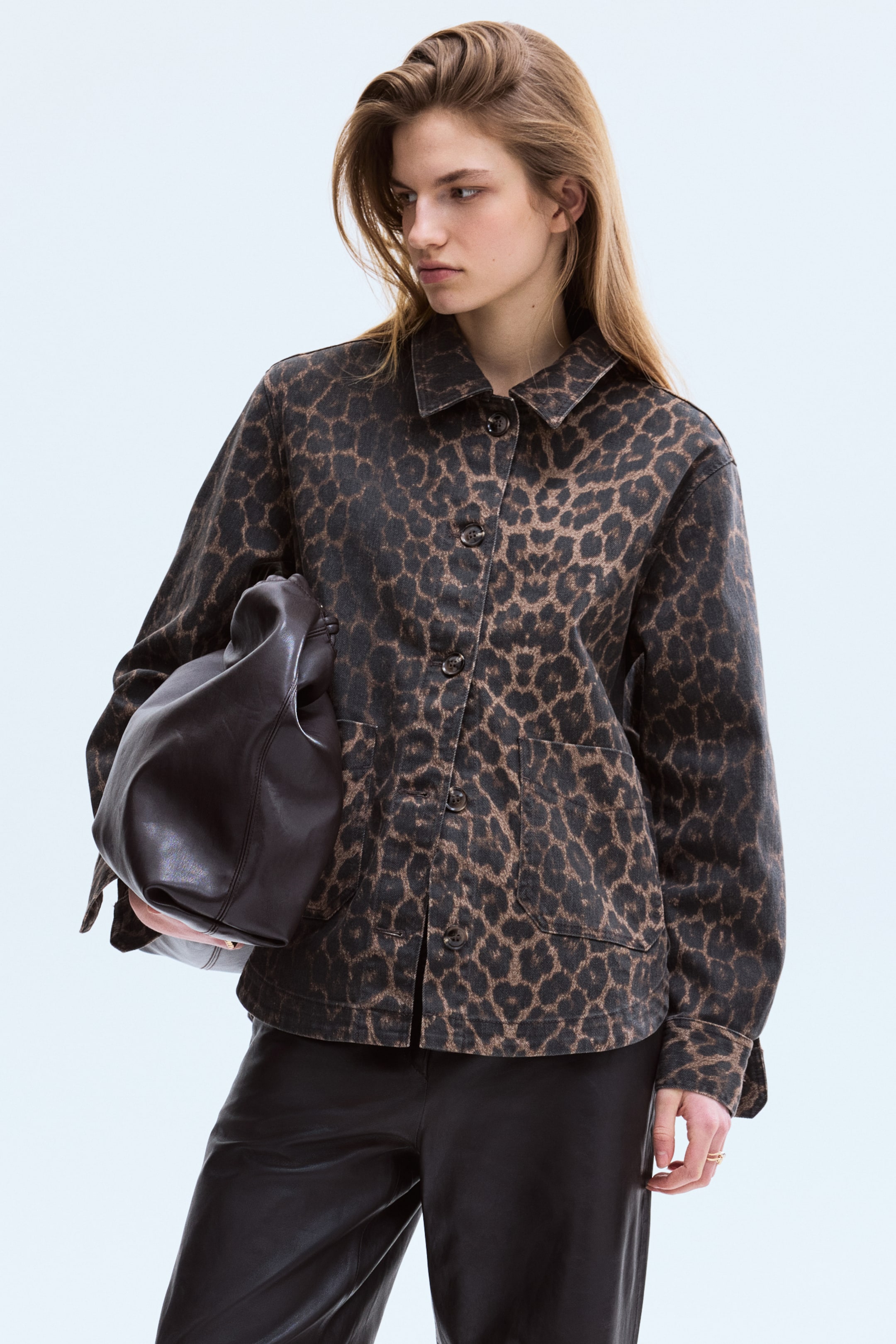 Agrandir l'image: La mannequin porte une surchemise à imprimé léopard, dotée d'un col, d'une fermeture boutonnée avec des boutons foncés, de deux poches plaquées à l'avant, d'épaules tombantes, de manches longues avec poignets boutonnés, et d'un ourlet droit. La mannequin tient un sac en cuir foncé devant la surchemise.