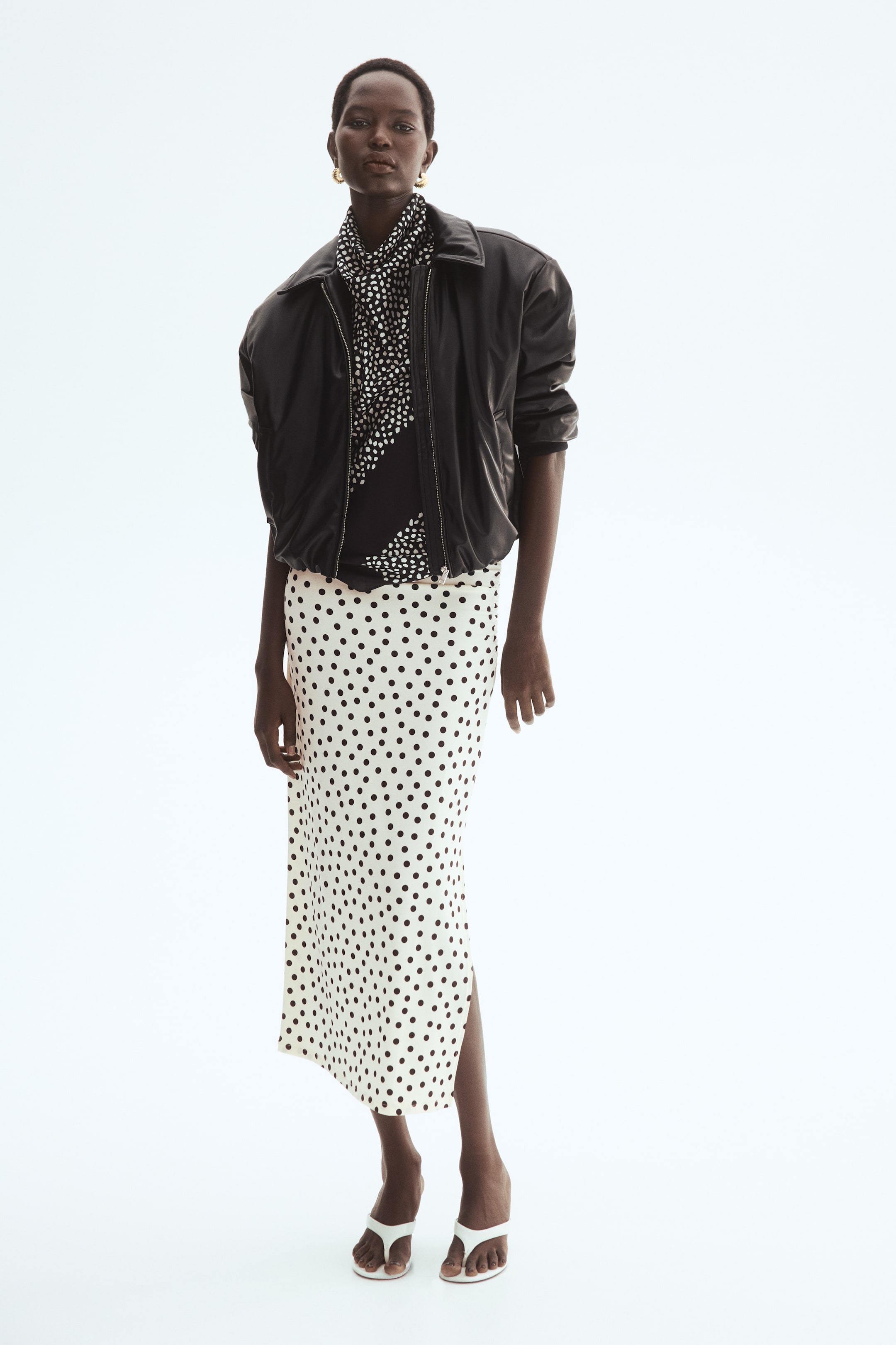 View larger image: The model is wearing a fitted midi skirt crafted from jersey, adorned with a black polka dot pattern set against a light backdrop. This skirt boasts a high waist, gracefully draping at the sides, and features a straight-cut hem with a high slit on one side, perfect for adding a touch of flair.