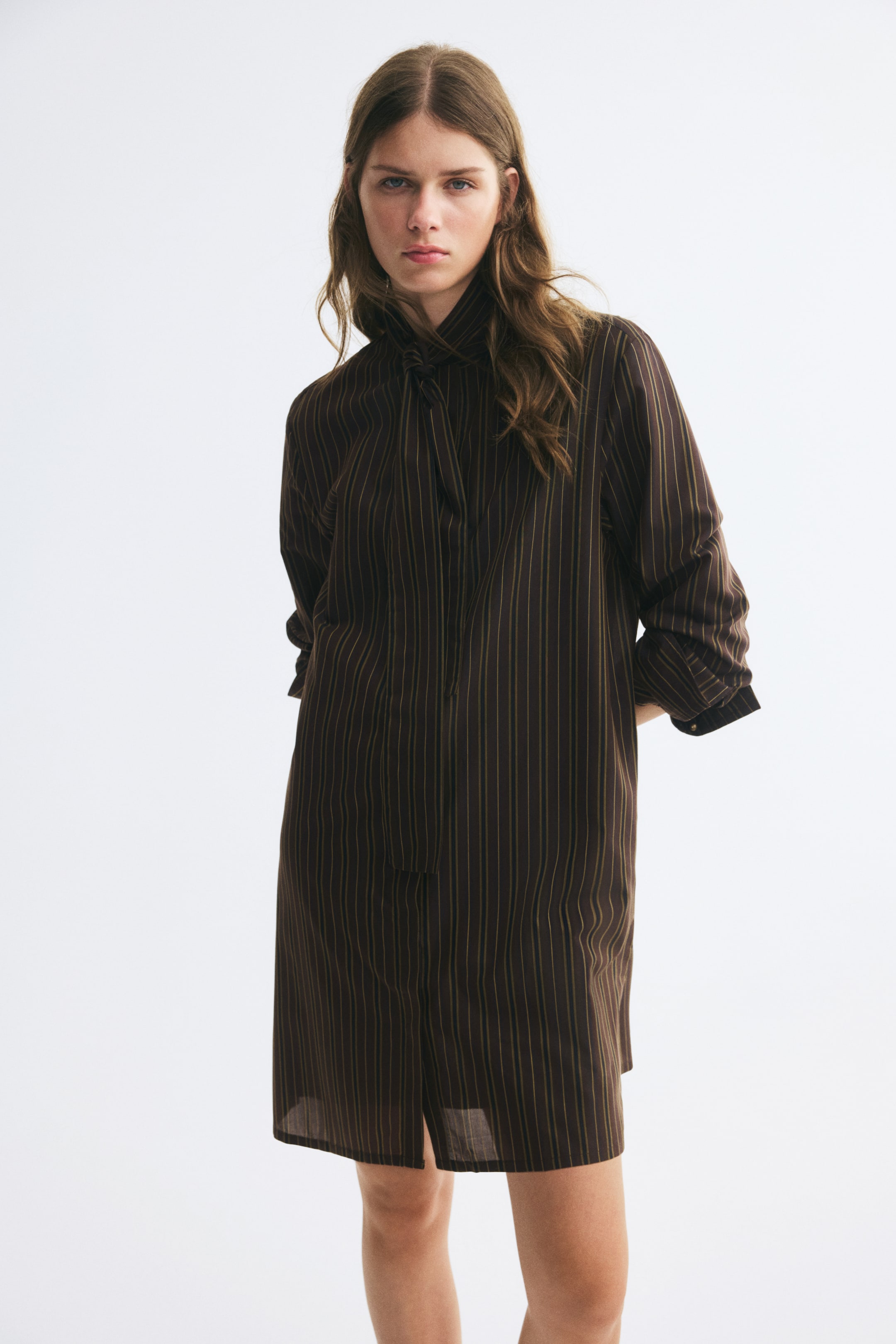 Voir une image plus grande: Une robe courte et surdimensionnée en tissu brun foncé à fines rayures verticales est portée par le mannequin. La robe présente un col montant avec une longue écharpe drapée, des manches longues avec poignets repliés et un ourlet droit.
