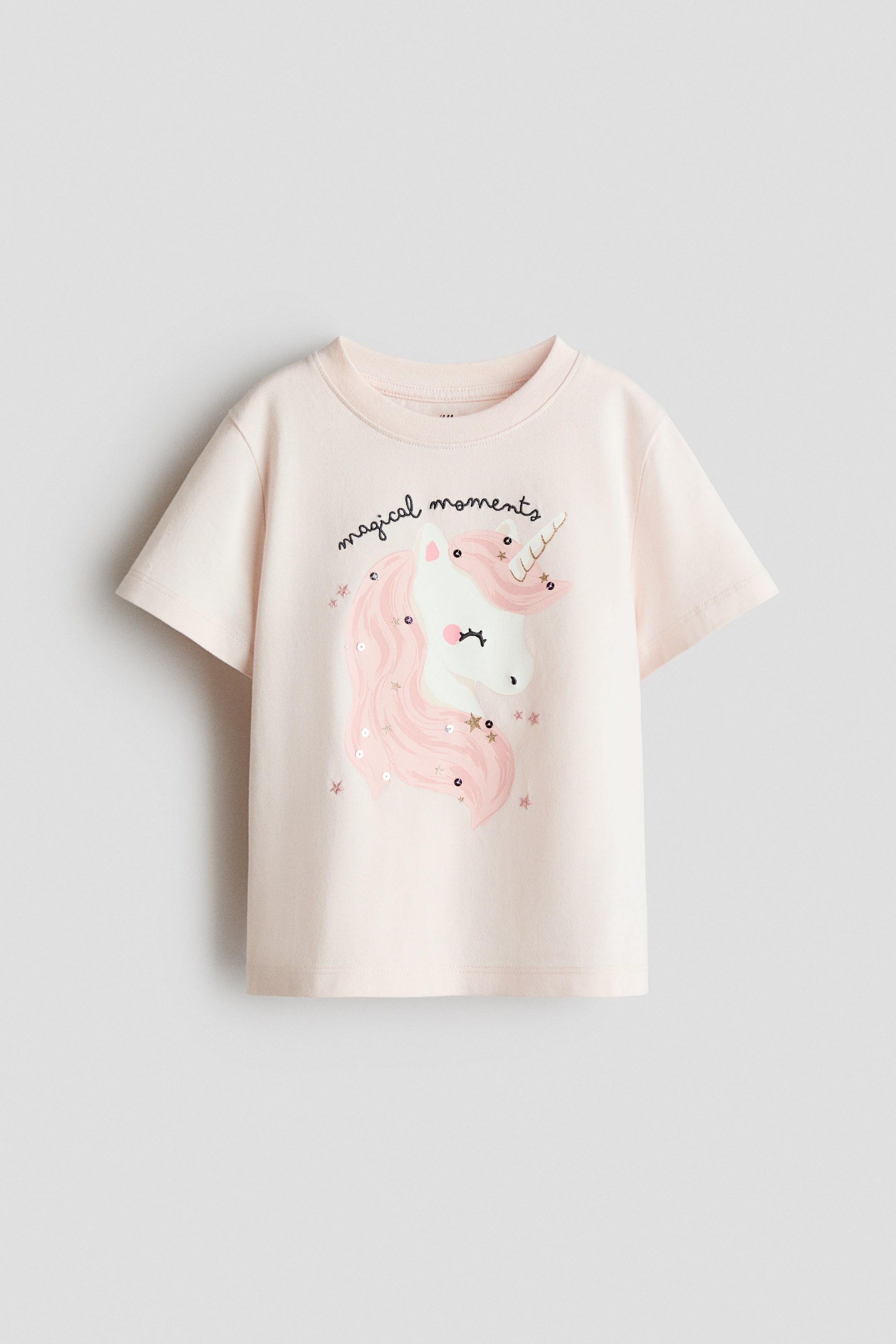Größeres Bild ansehen: Ein hellrosa T-Shirt zeigt seine VORDERSEITE, mit einem runden, gerippten Halsausschnitt und kurzen Ärmeln. Ein Einhornkopf-Grafik schmückt die Vorderseite, mit rosa Pailletten und goldenen Sternen in der Mähne, darüber steht der Text "magical moments".